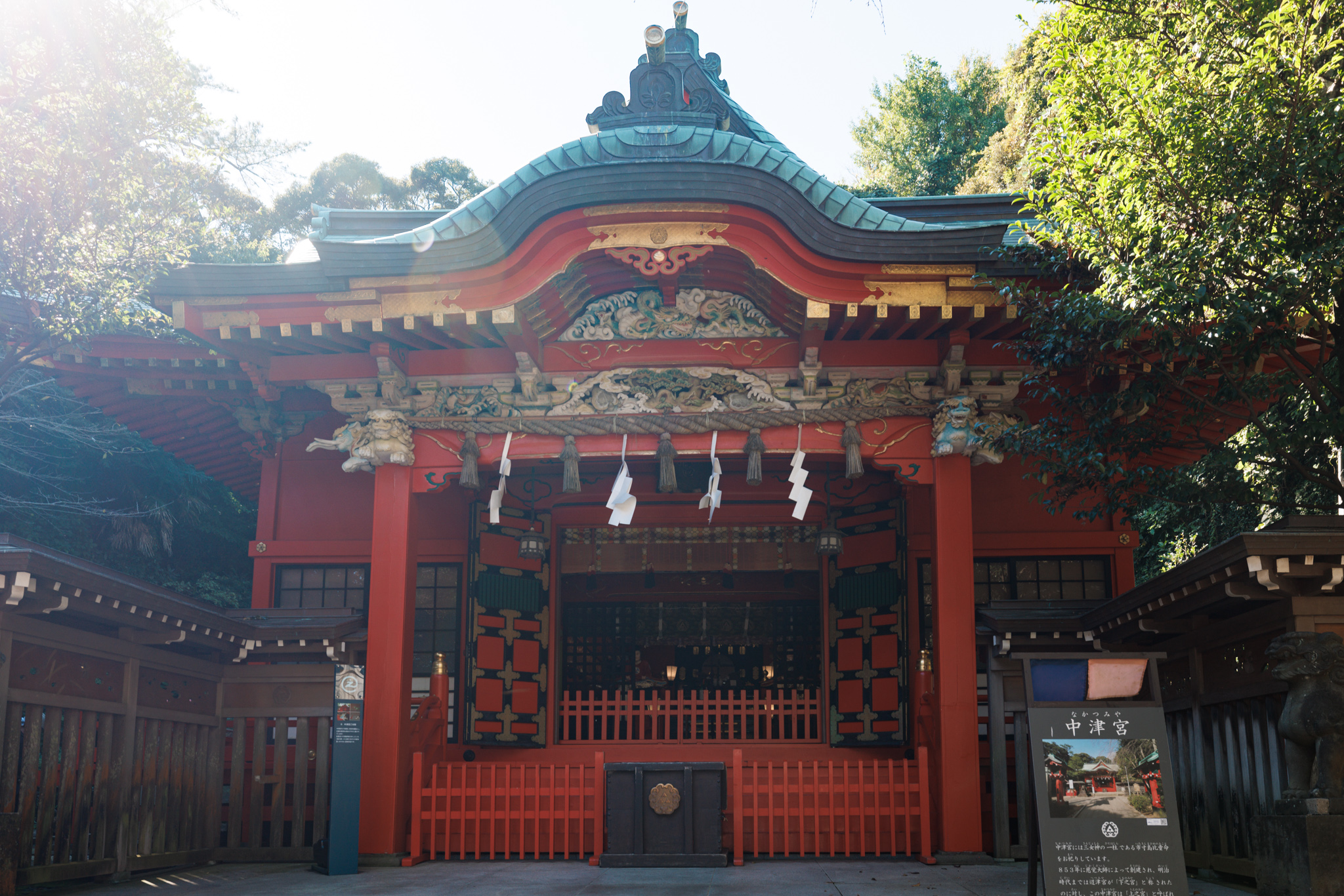 The front of a vermilion shrine with a green copper roof, gold and painted carvings under the eaves, white paper streamers and a low red fence, trees behind and a signboard to the right