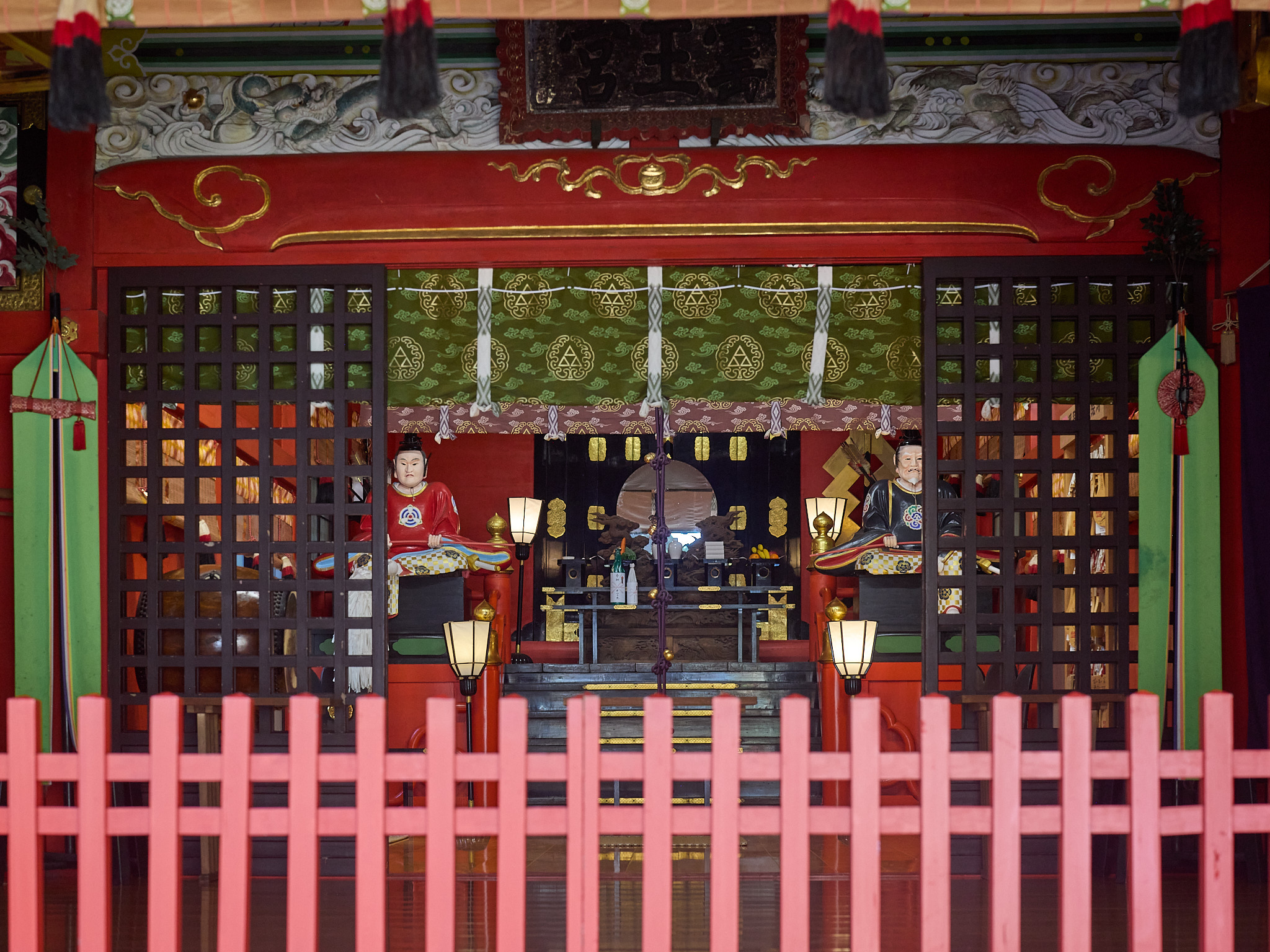 Inside a red shrine hall behind a low red fence and lattice doors: two seated figures in red robes on either side, lanterns and a round mirror at the back