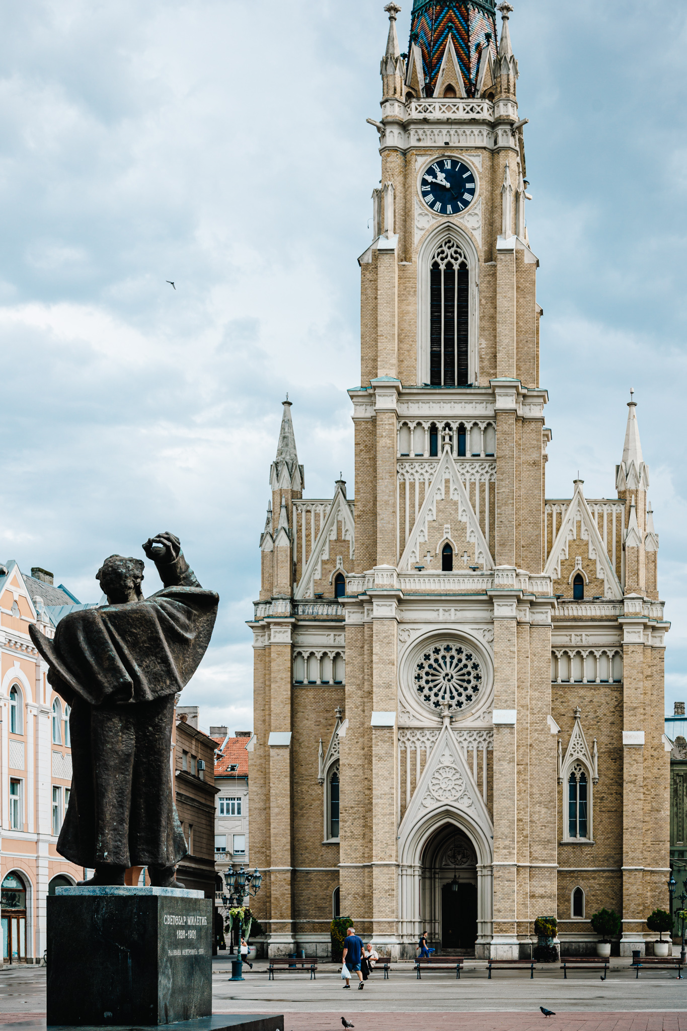 The bronze Svetozar Miletić monument seen from behind on Freedom Square, with the Name of Mary Church beyond.