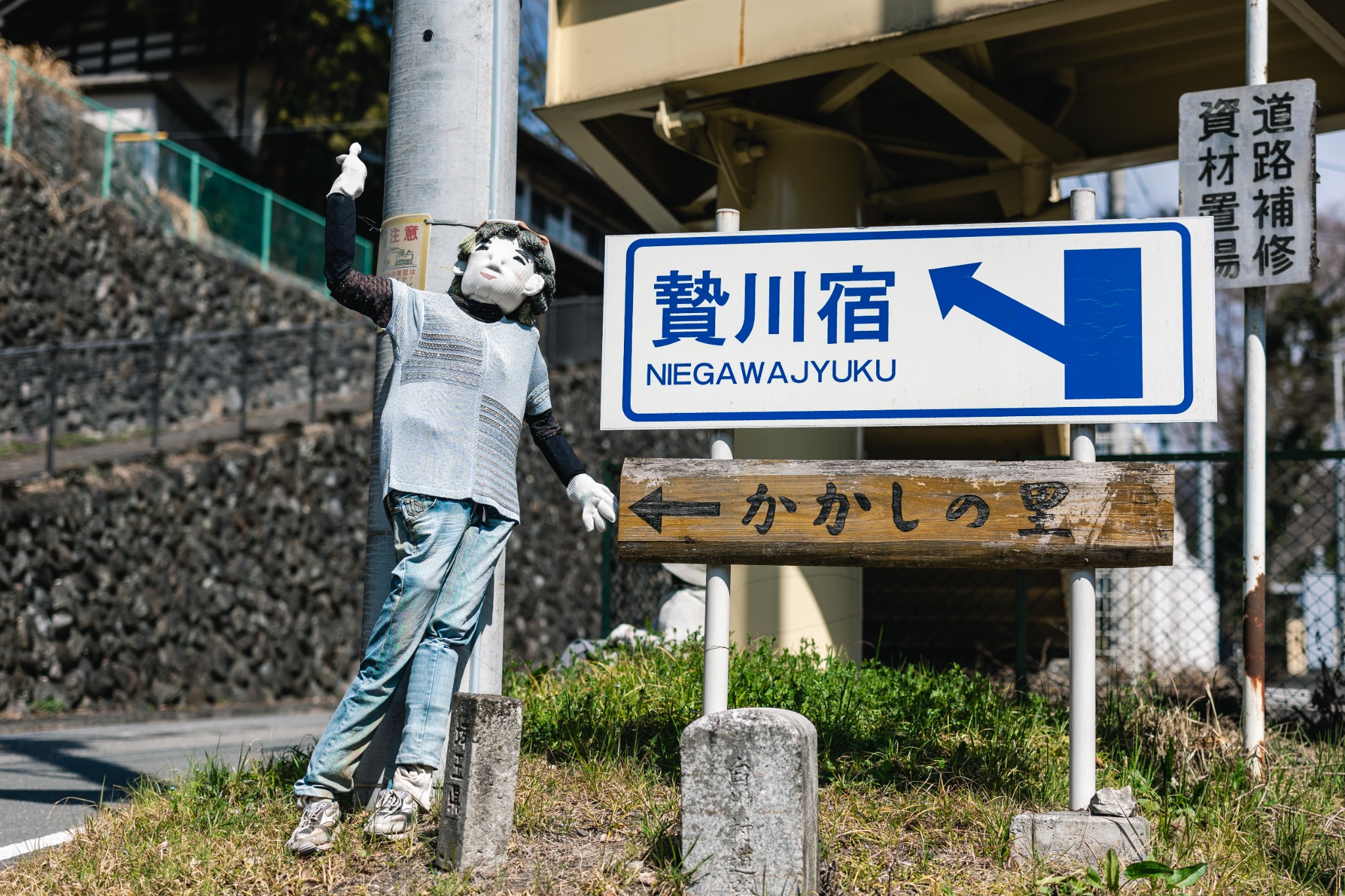 A handmade scarecrow raises a gloved hand beside the Niegawa-juku sign in Chichibu.