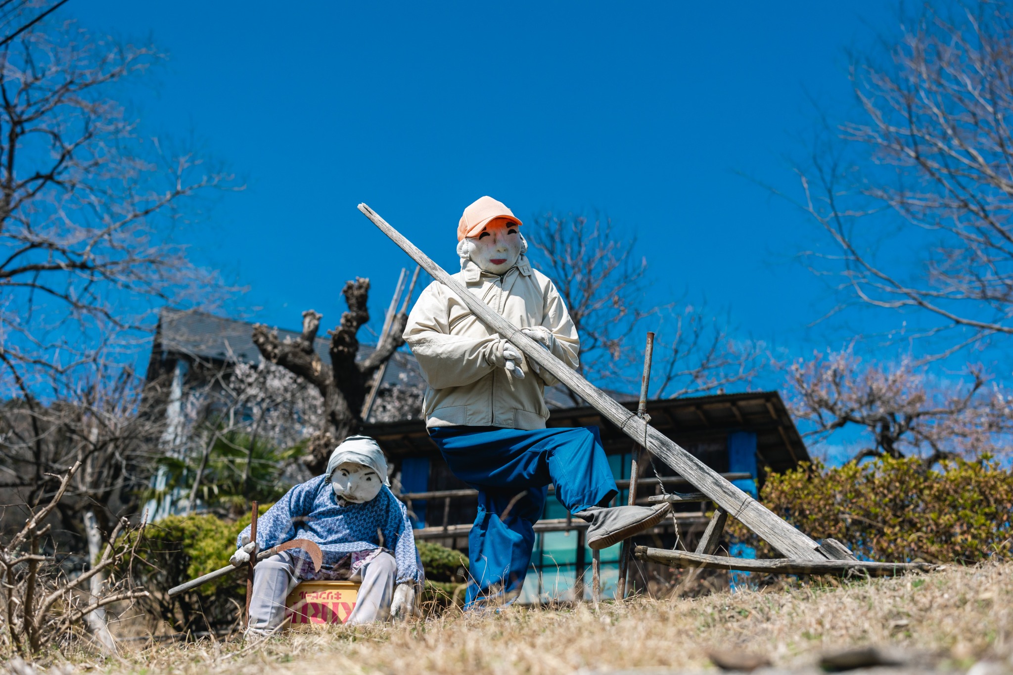 A handmade scarecrow in Niegawa-juku raises one leg while holding a long wooden tool.