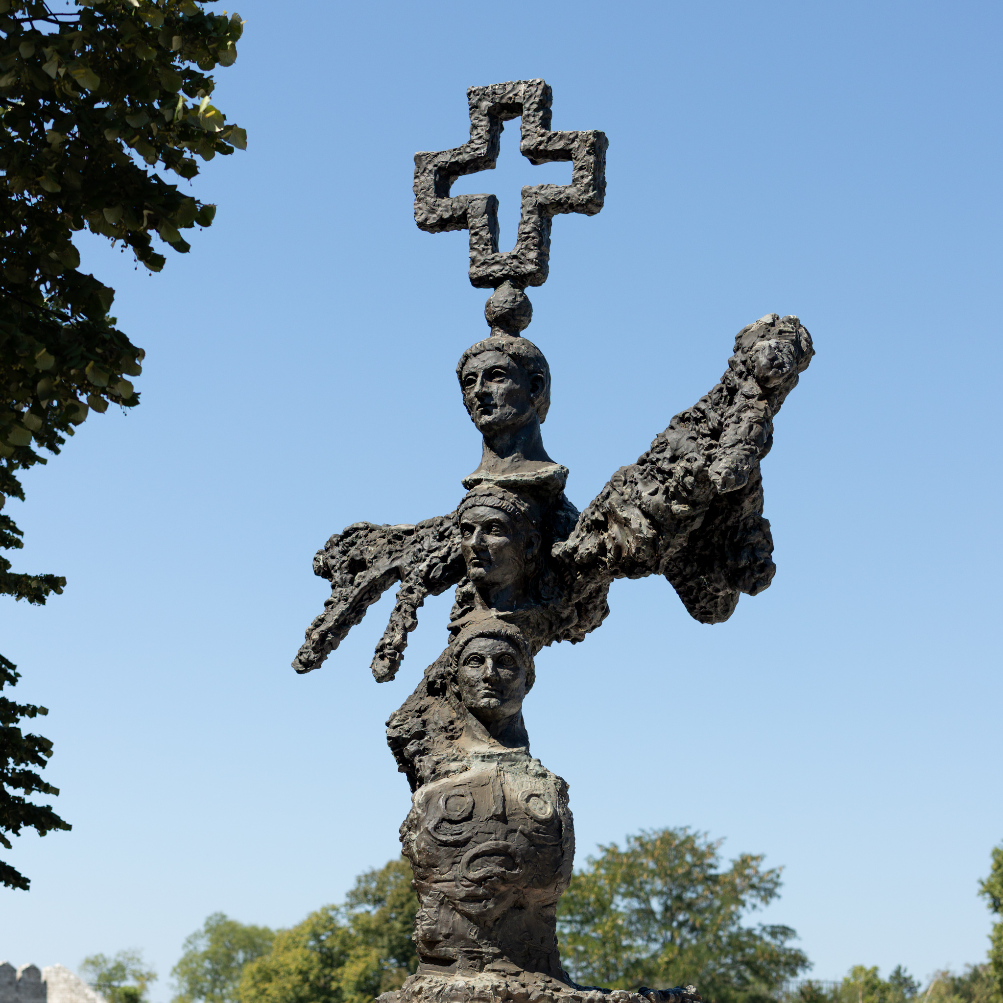 The Metamorphosis Constantiniana sculpture against a blue sky: a spiral-patterned base, three stacked portraits of Constantine, a pair of rough outspread wings and a cross on top