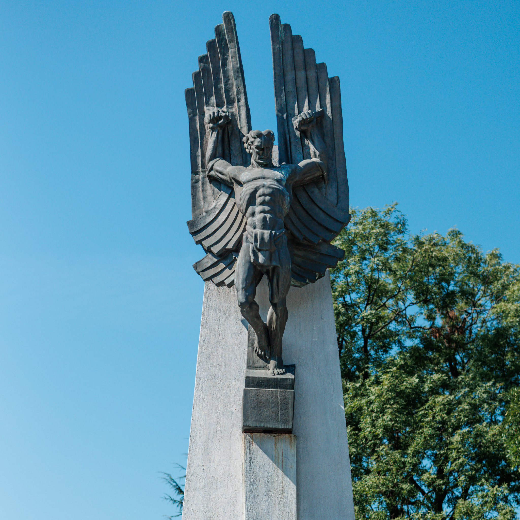 Close view of the bronze Icarus gripping a tall wing in each raised fist, one foot on a small block and the other leg bent and lifted, against a blue sky