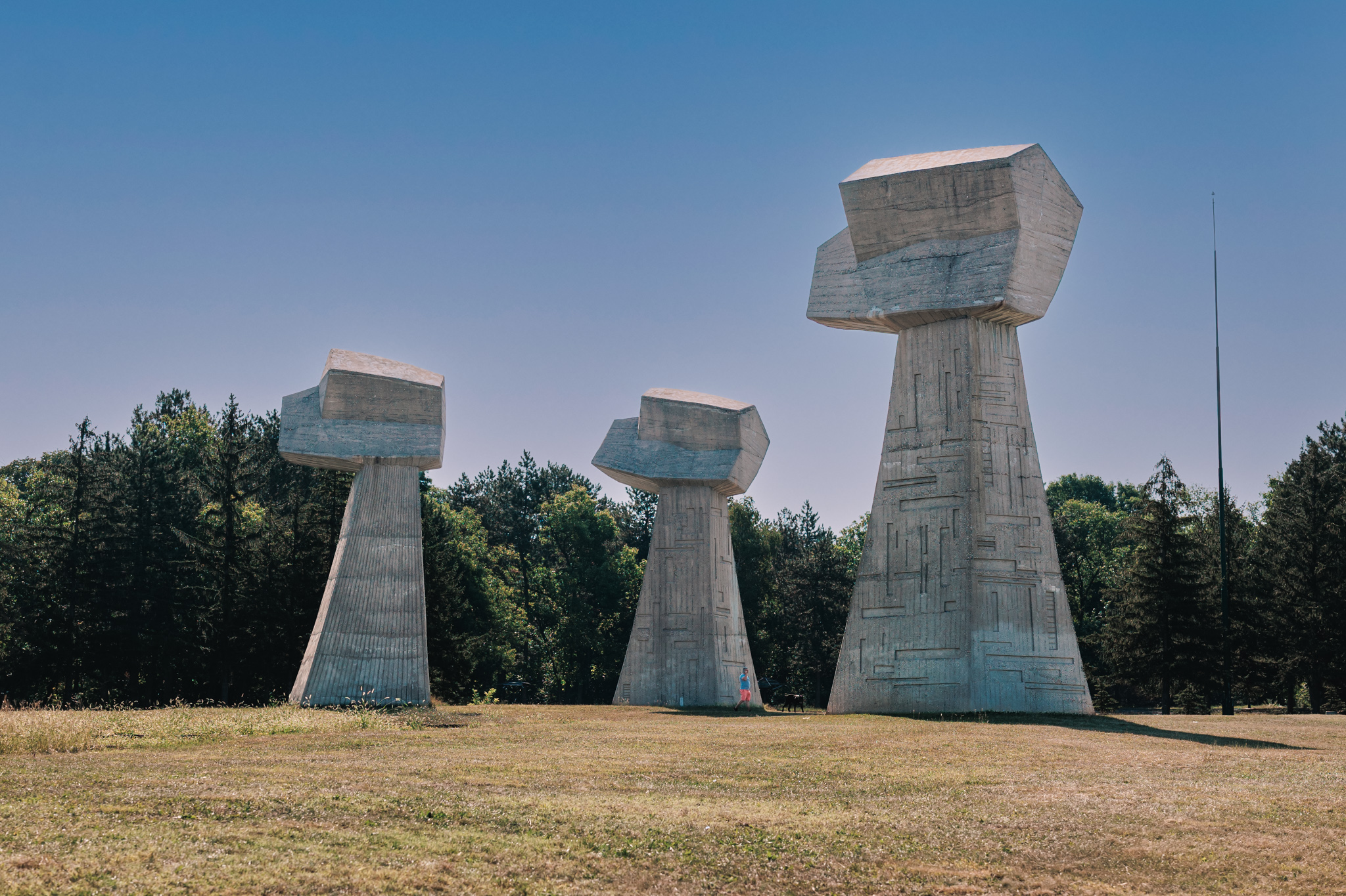 Three tall concrete fists of different sizes rising from a mown lawn at Bubanj Memorial Park in Niš, with pine trees behind