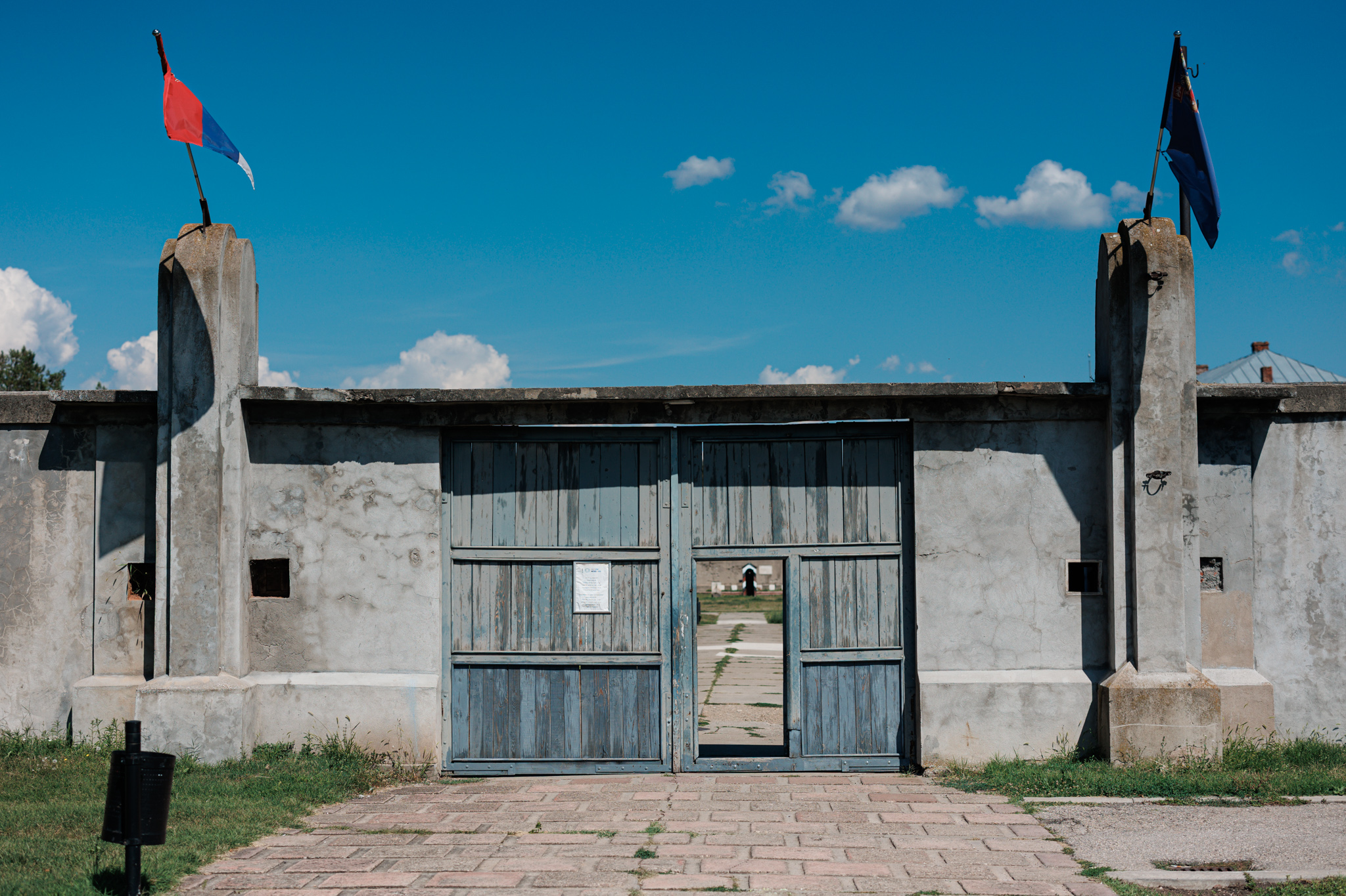 The weathered blue wooden gate in the grey wall of the former Crveni Krst camp in Niš, with flags above the gateposts and a glimpse of the yard through the gap