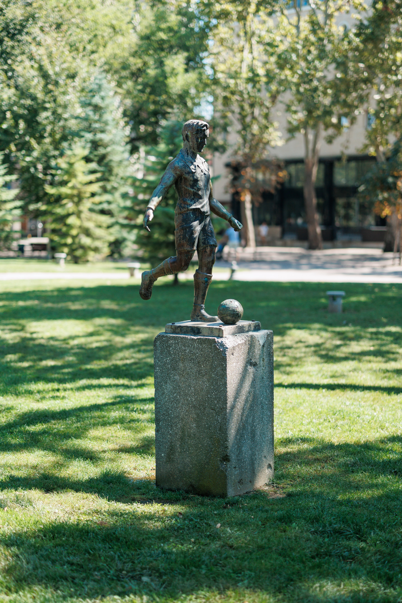 Bronze statue of a footballer in shorts and socks about to kick a ball, on a small stone pedestal on a sunlit lawn