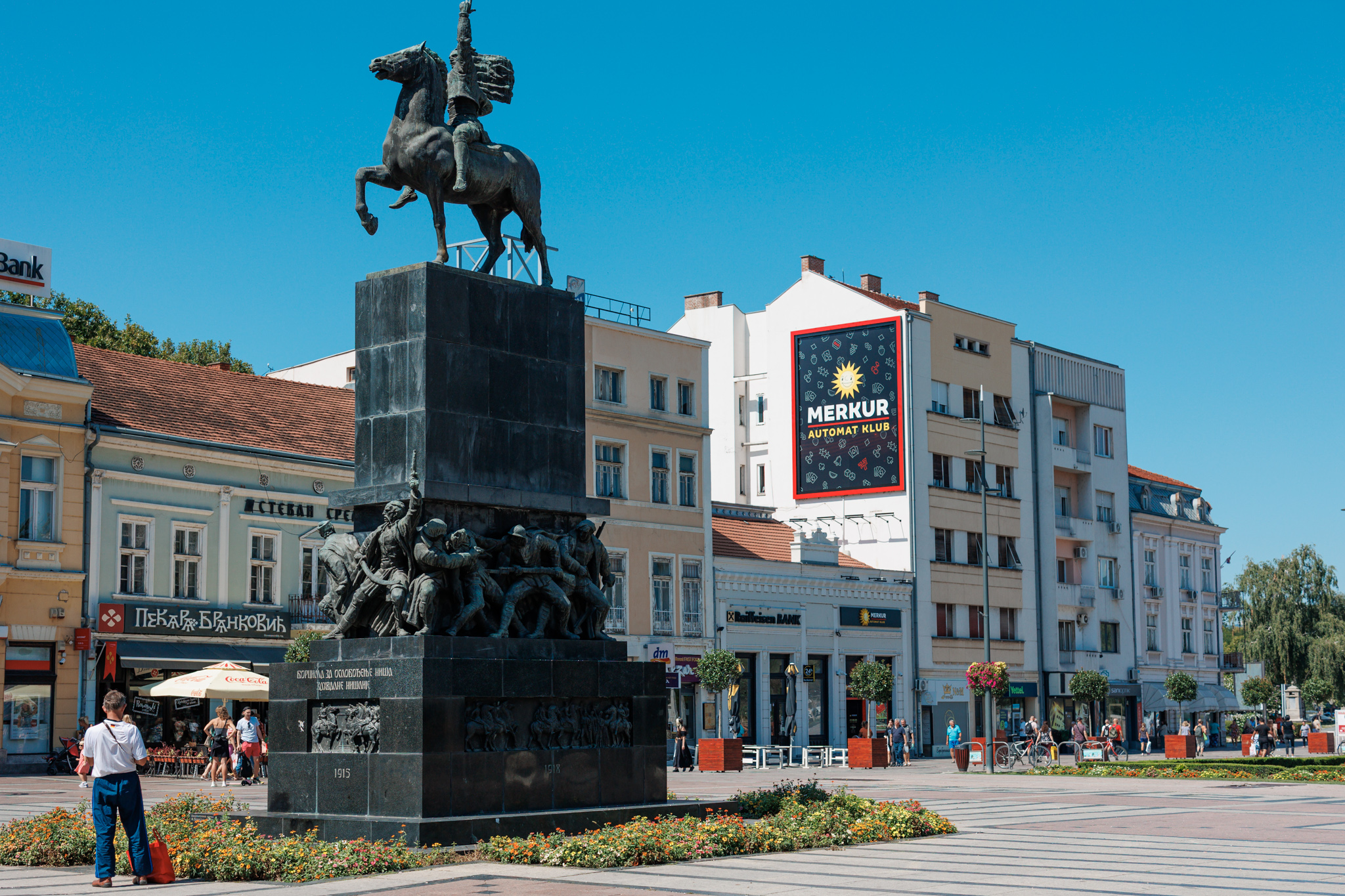 The Monument to the Liberators of Niš on King Milan Square: a bronze rider with a flag on a rearing horse above a crowd of bronze fighters, with a casino advertisement on a building behind