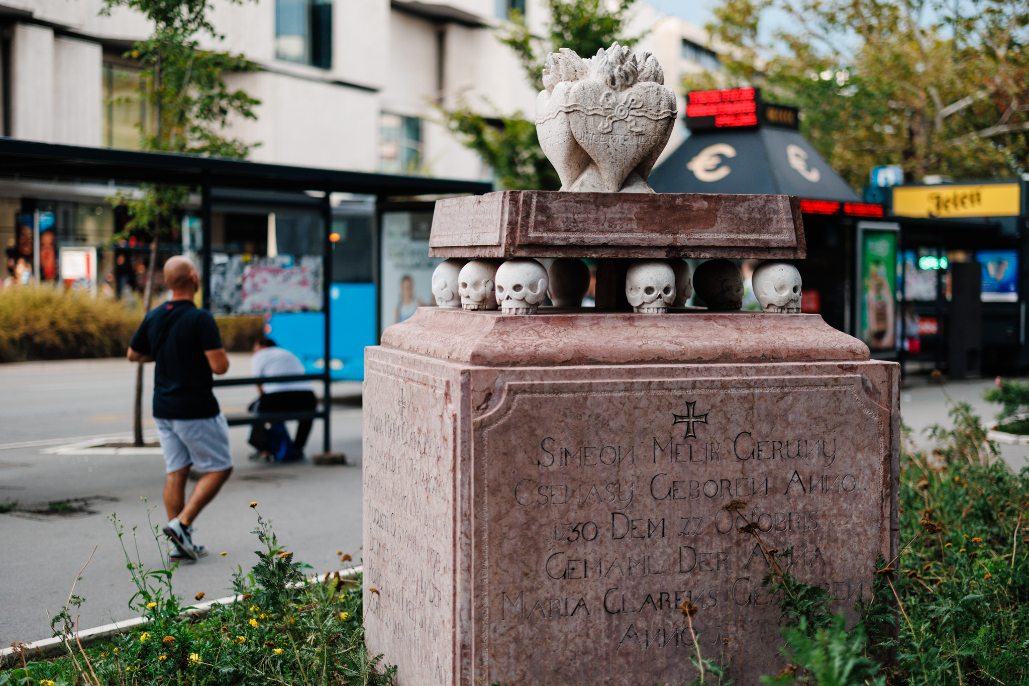 A pink marble Armenian family tomb beside a bus stop in central Novi Sad, with white skulls and intertwined hearts above it.