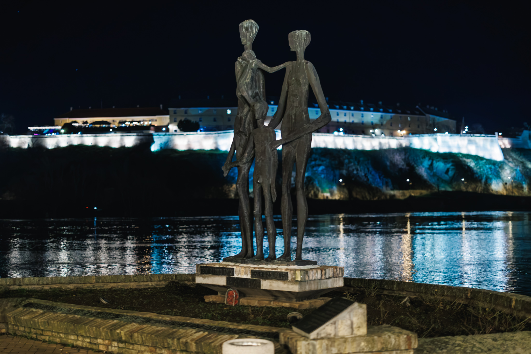 The Family sculpture at Novi Sad’s Raid Memorial beside the Danube, with Petrovaradin Fortress illuminated across the river.