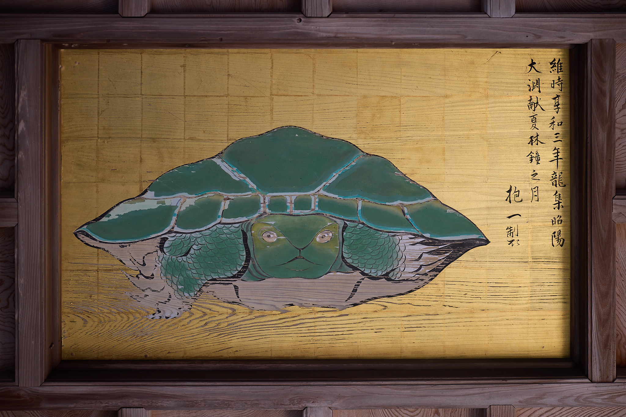 A painted turtle with a green shell and a face looking straight out, on a gold-leaf panel set in a wooden coffered ceiling, with lines of black brush inscription on the right