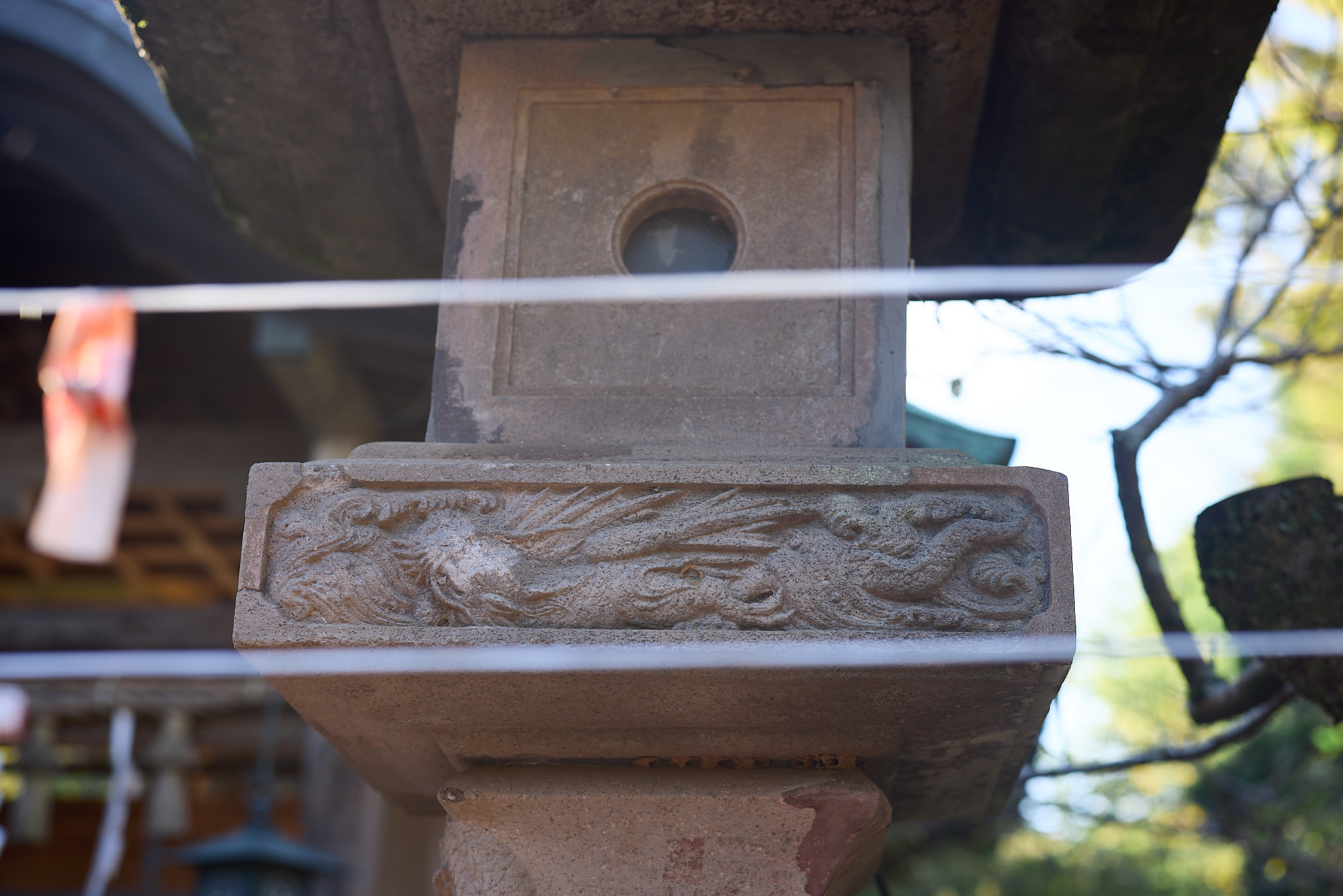 A carved band on a stone lantern showing a bird with swept-back feathers and a long tail, under the round window of the light box, with string lines crossing in front