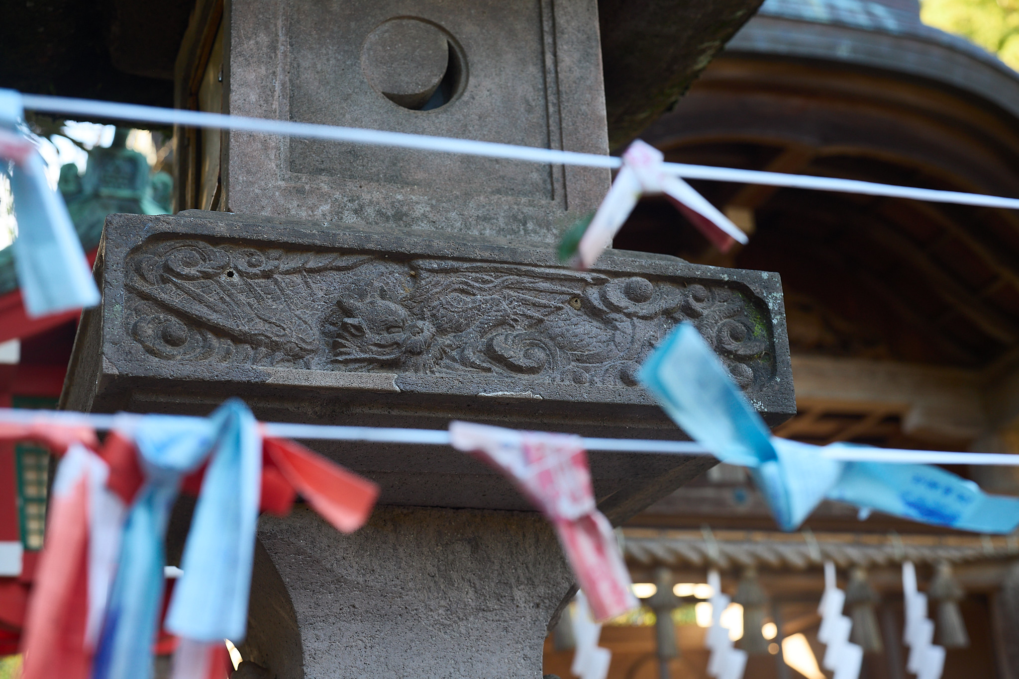 The top of a stone lantern with a crescent-shaped opening above a carved band, with red, blue and white paper strips tied on lines in front
