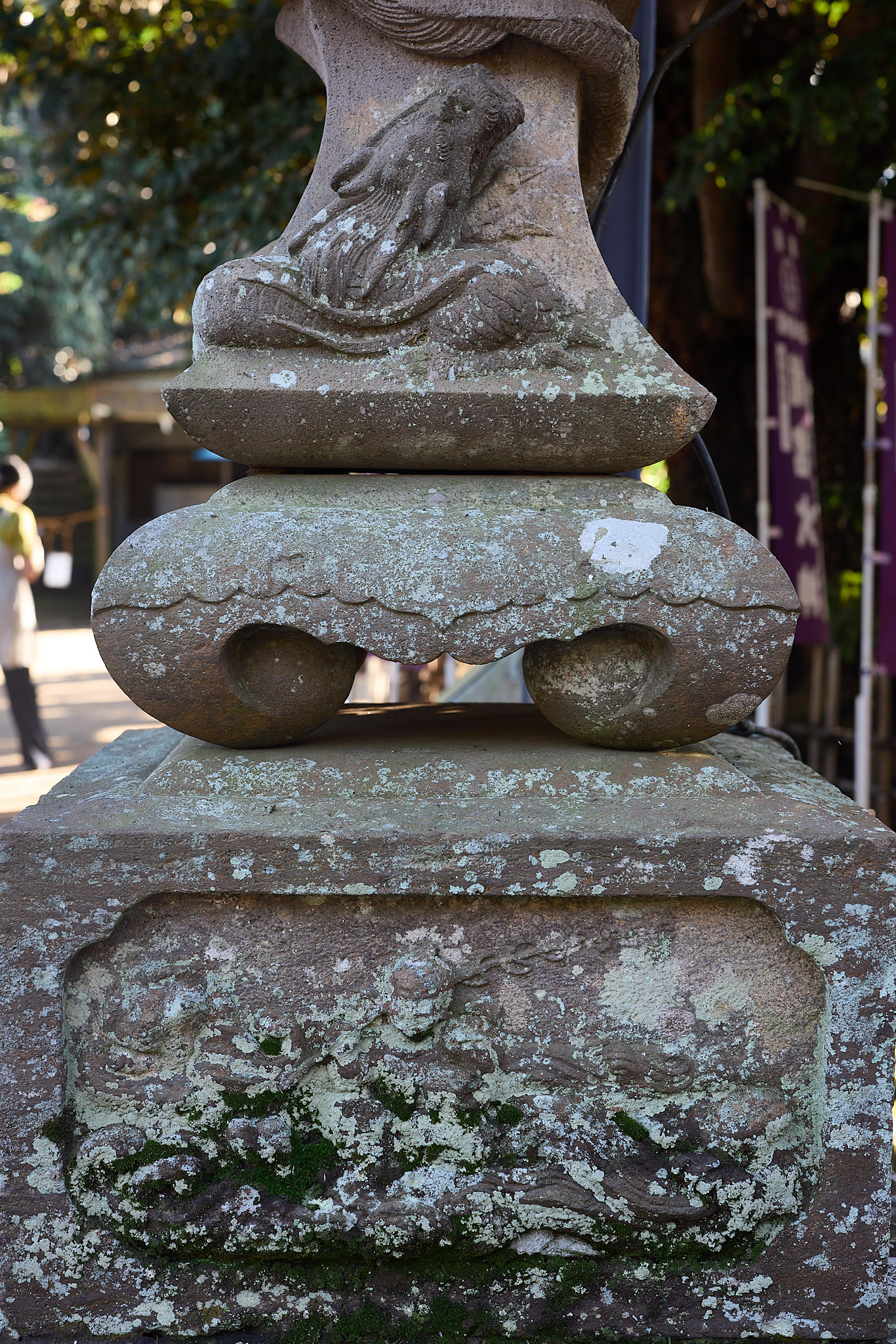 The lower part of a stone lantern with a fish carved climbing the shaft and a relief on the base almost hidden under pale lichen and moss