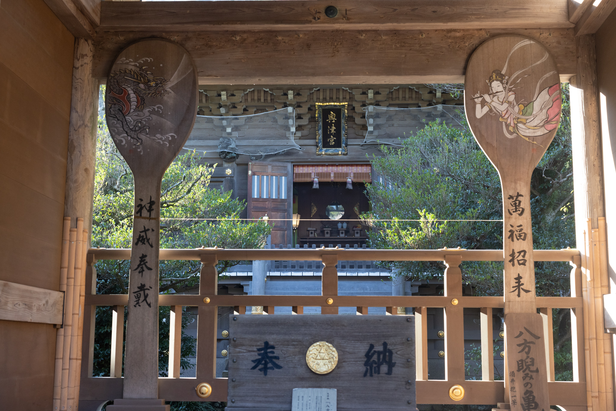 Two giant wooden rice paddles on either side of a view to the shrine building, one painted with a dragon in waves and the other with a heavenly maiden playing a flute, each with four large characters on its handle