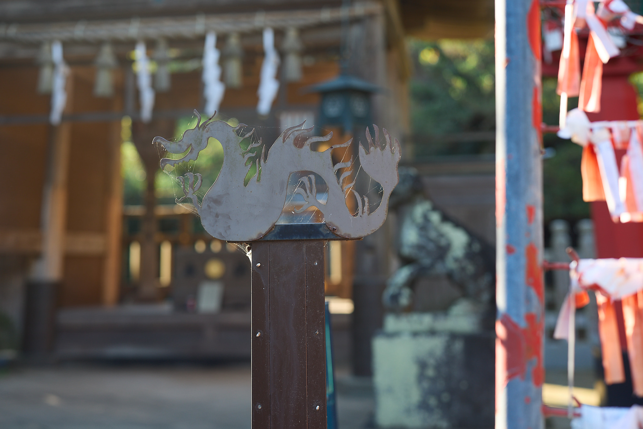 A flat dragon cut from sheet metal on top of a wooden post, cobwebs strung between its claws, with a blurred shrine building and stone guardian lion behind