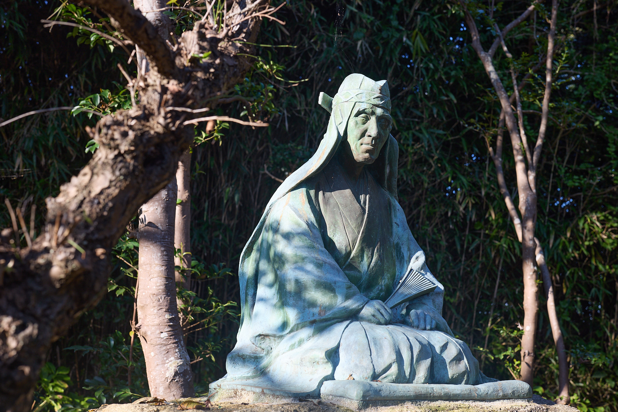 A seated bronze statue of a man in a hood and robes holding a folding fan, green with age, among trees in dappled sunlight