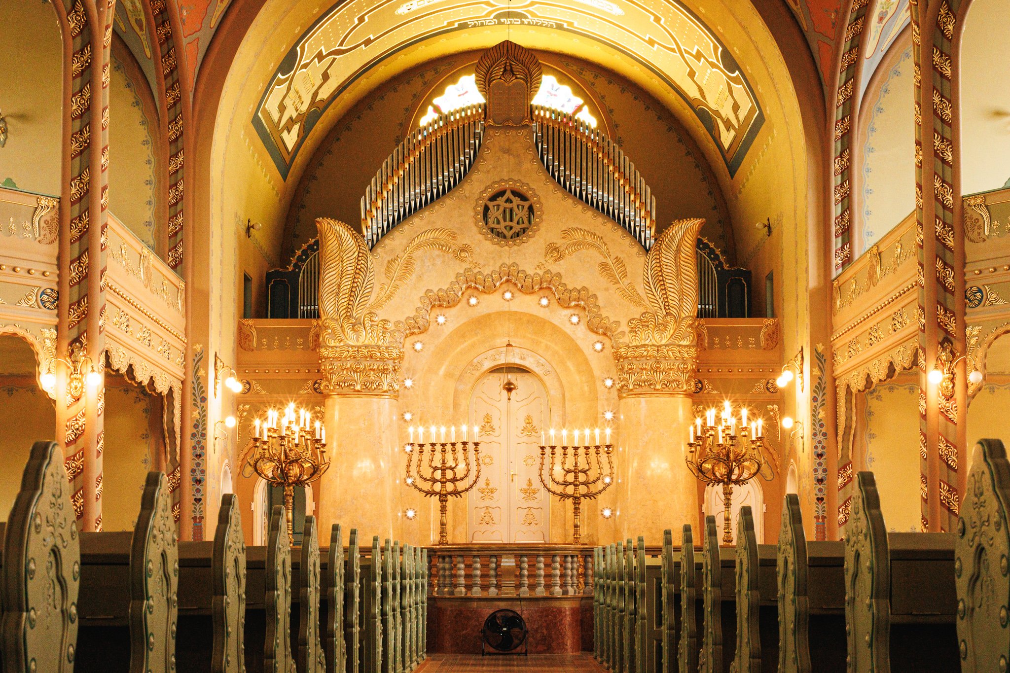 The gilded Torah ark of Subotica Synagogue beneath organ pipes, flanked by candelabra, with rows of green benches in front and a black electric fan on the floor