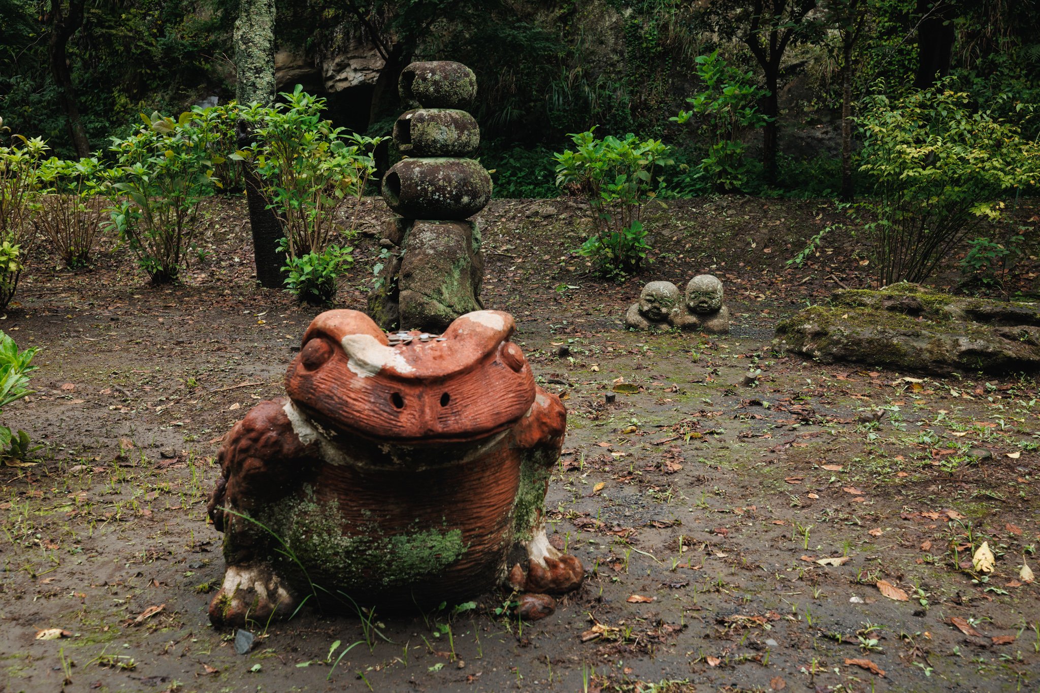 A red frog among small stone figures on the path at Kenchōji Temple, with coins resting on its head.