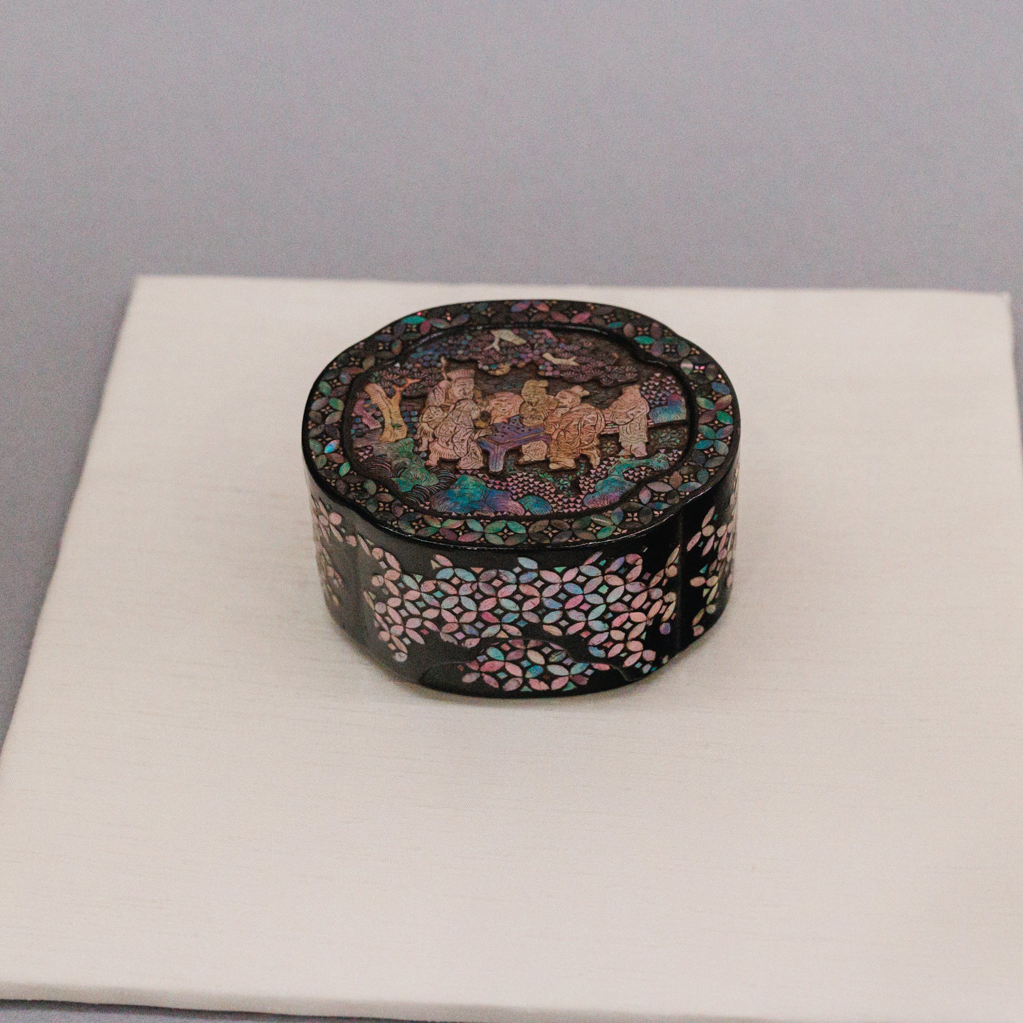 A small black lacquer box covered in iridescent mother-of-pearl, with figures around a table on the lid and flower patterns on the sides