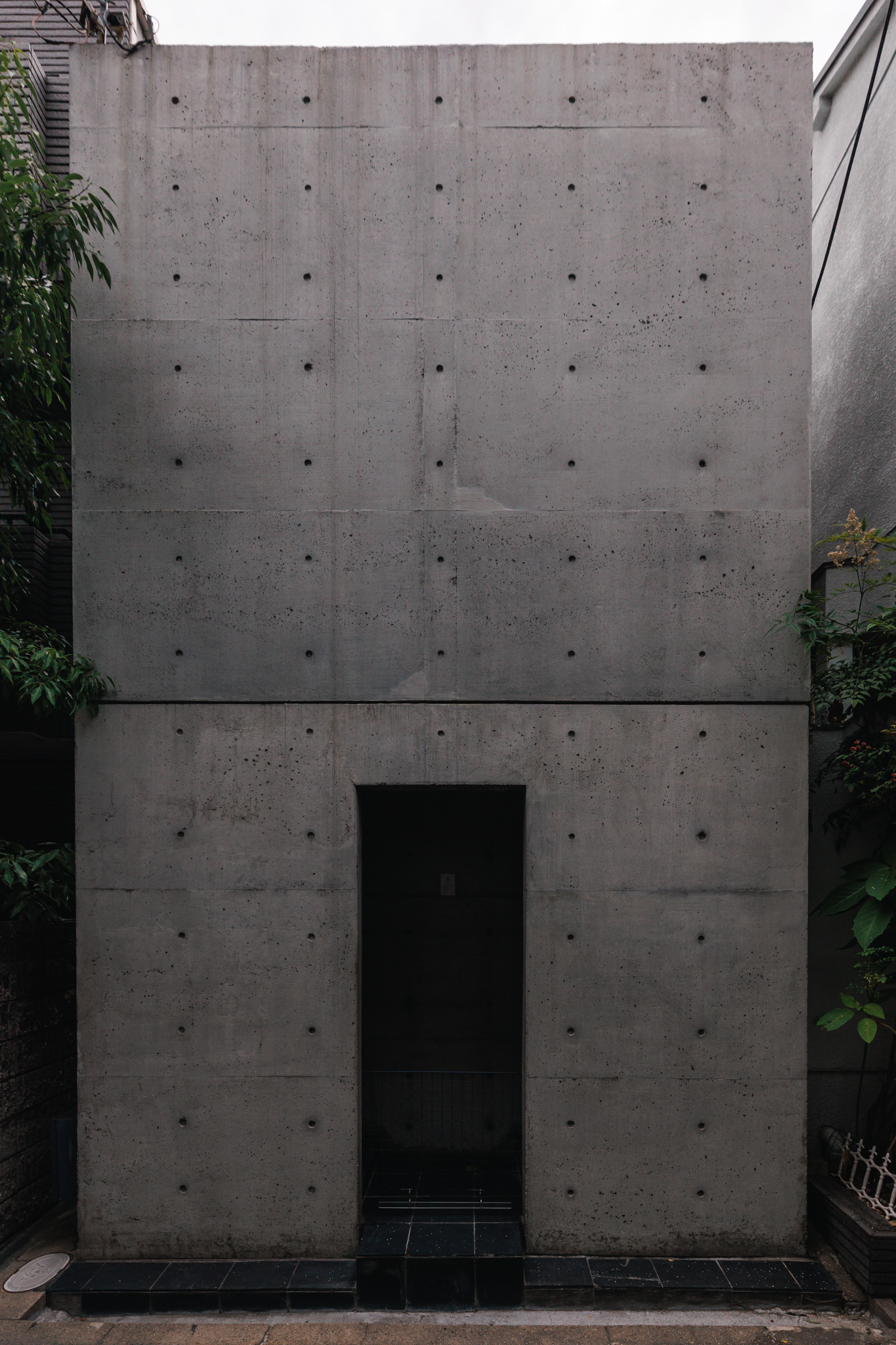 The windowless exposed-concrete façade of the Row House in Sumiyoshi, two storeys high with a single recessed doorway, between trees and neighbouring houses