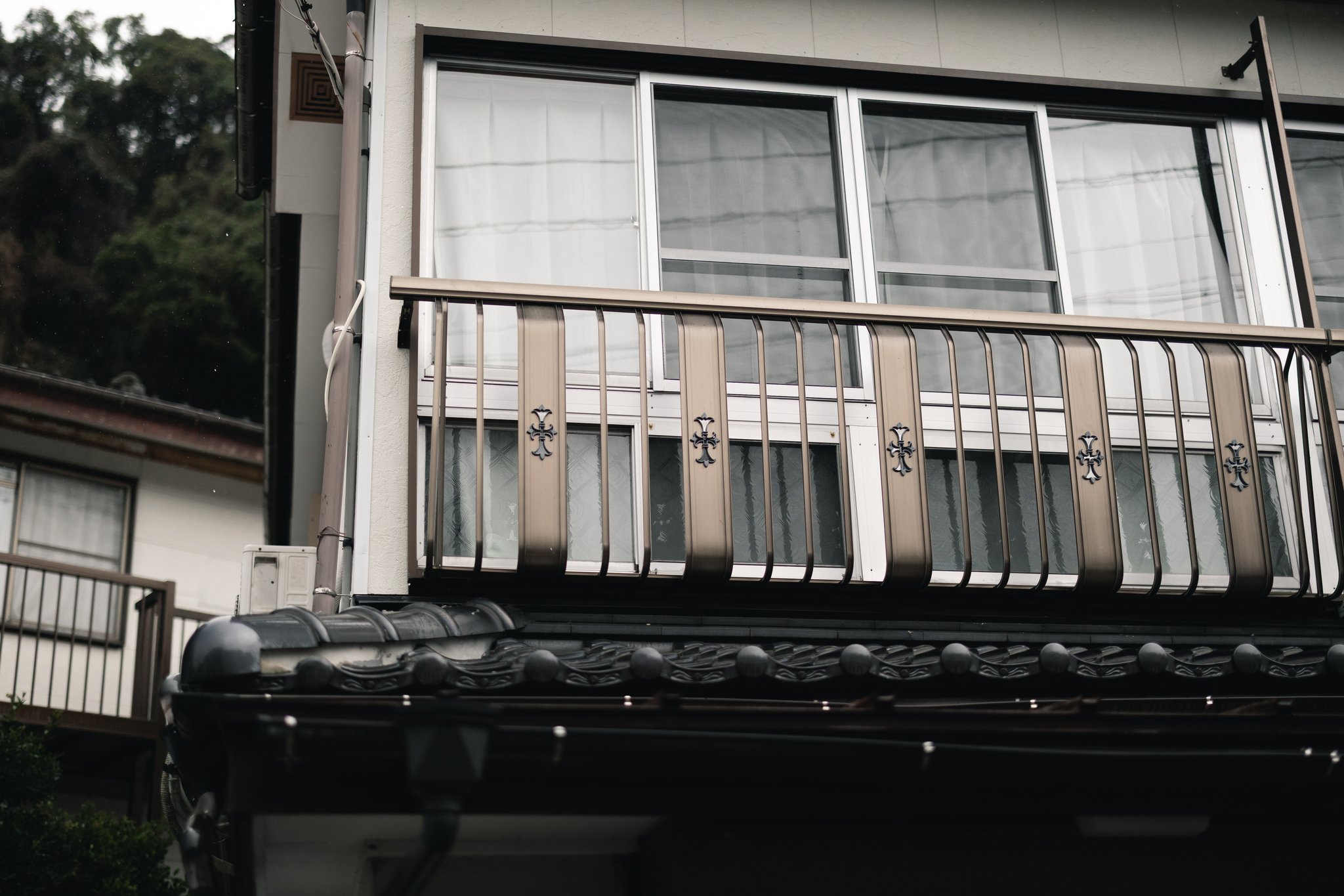 A first-floor balcony in Sakitsu with metal railings decorated with crosses, above a Japanese tiled roof