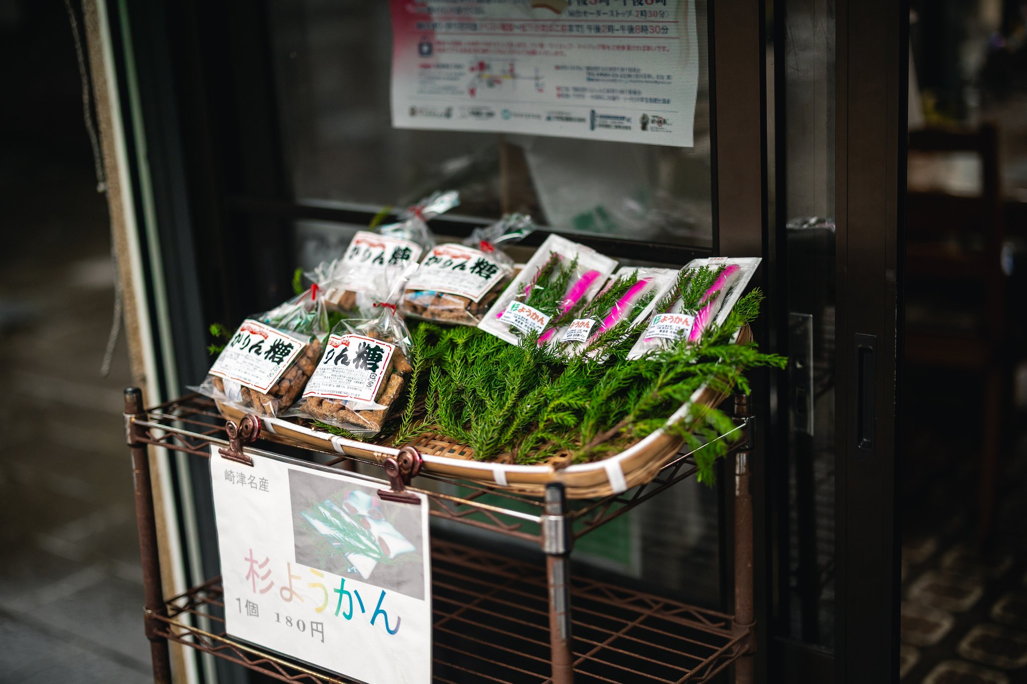 Packs of sugi yōkan on cedar leaves and bags of karintō in a basket outside a shop, with a 180-yen sign