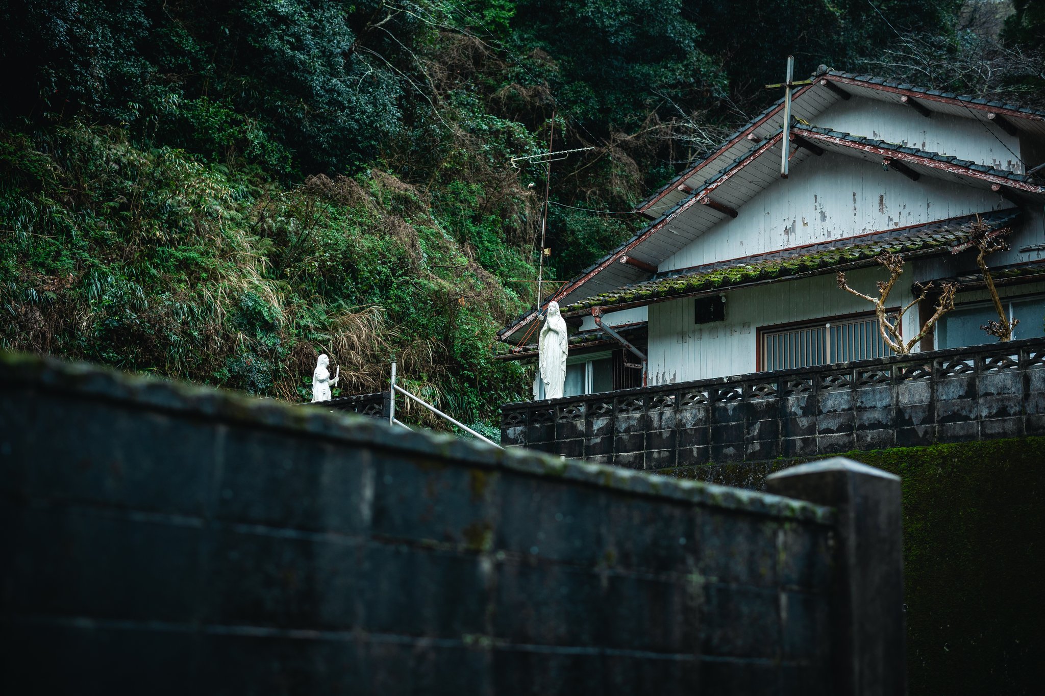 An old white wooden building with a wooden cross on its gable beside a Japanese roof ornament, and two white statues of Mary on the wall below