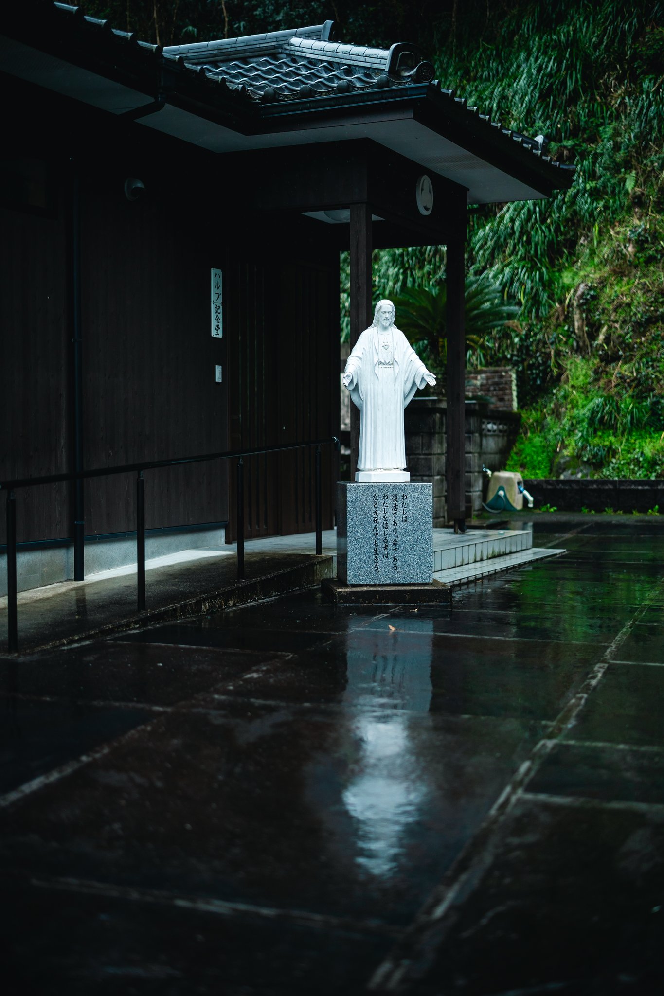 A white statue of Christ on a granite plinth inscribed with a verse from John, outside the Halbout memorial hall, on wet paving