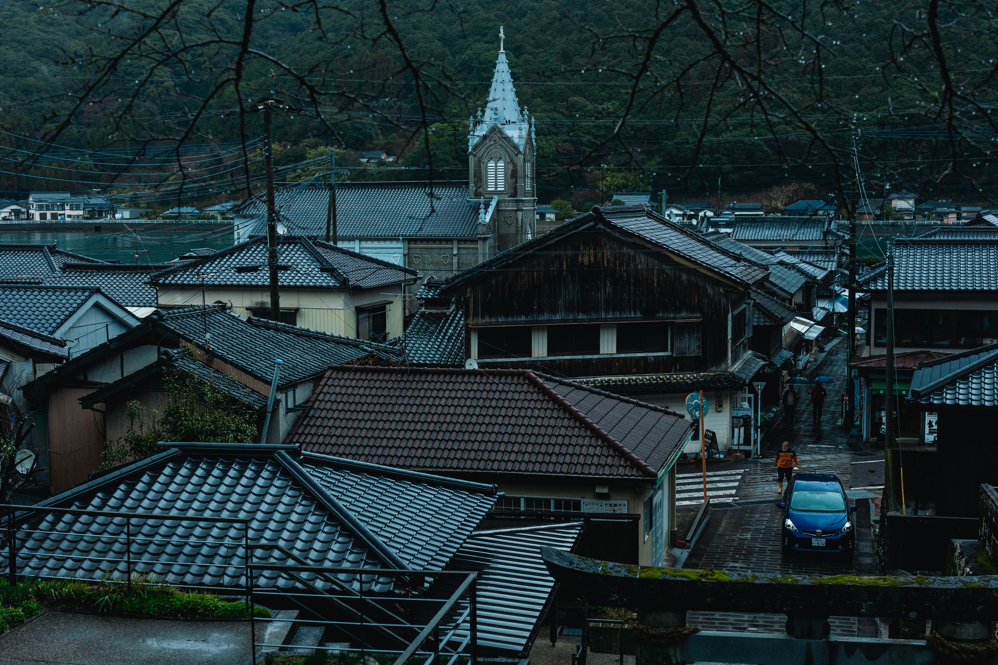 Looking down from the shrine steps over the tiled roofs of Sakitsu to the church spire and the inlet, with people under umbrellas in the lane