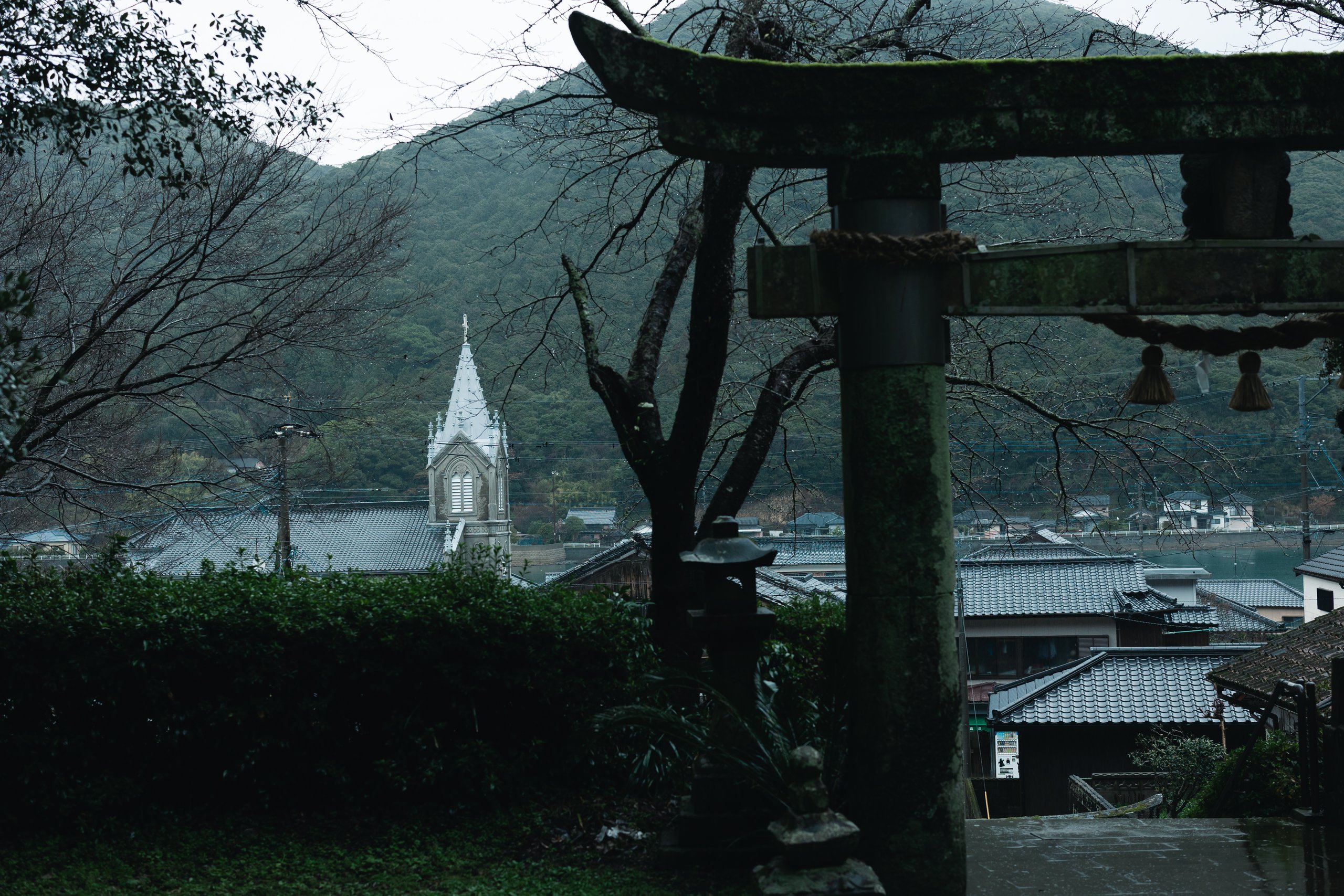 The Gothic spire of Sakitsu Church rising above tiled roofs, seen past the mossy stone torii of Sakitsu Suwa Shrine on a rainy day