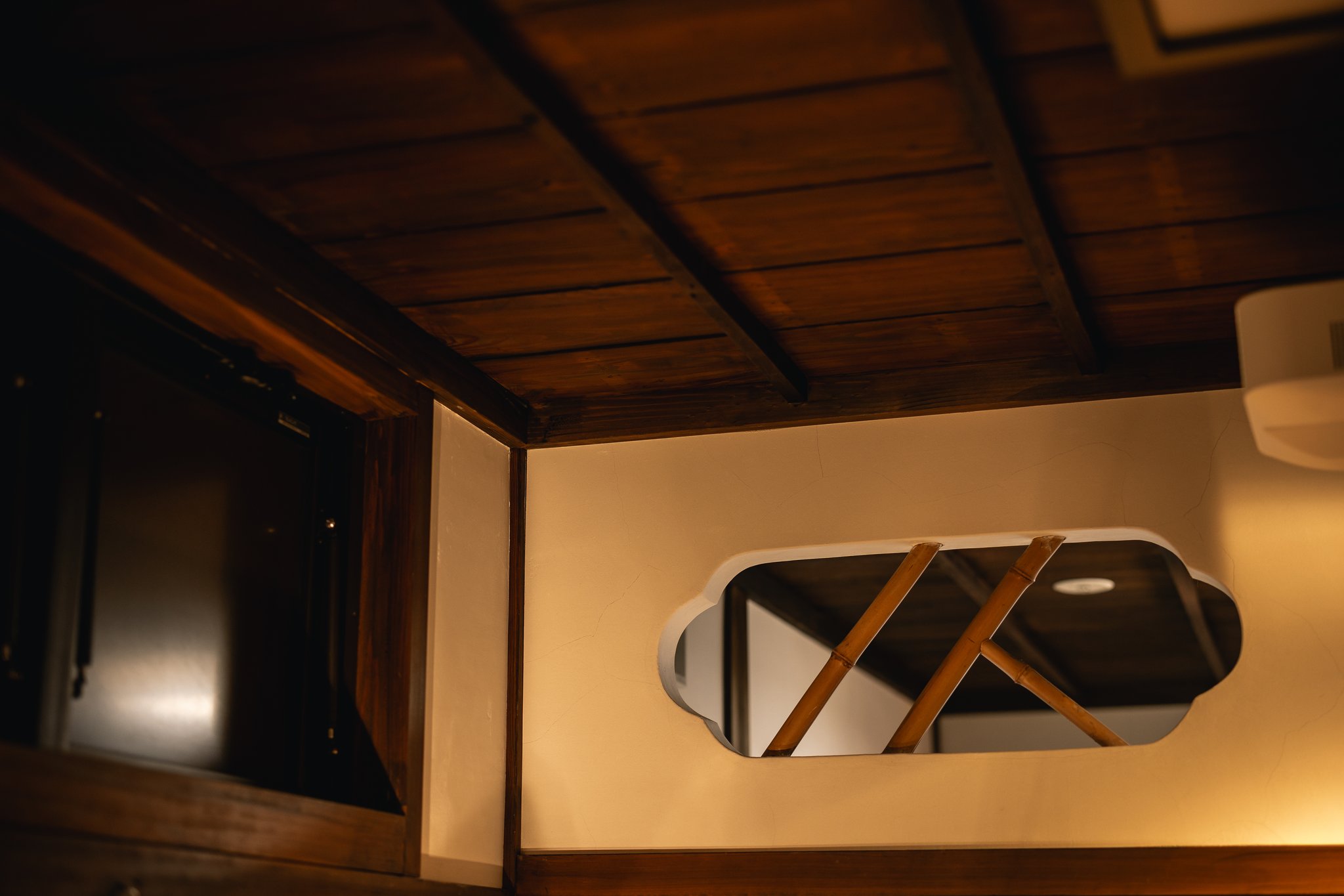 A dark wooden ceiling and a plaster wall with a rounded window crossed by diagonal bamboo, inside Minato-ya