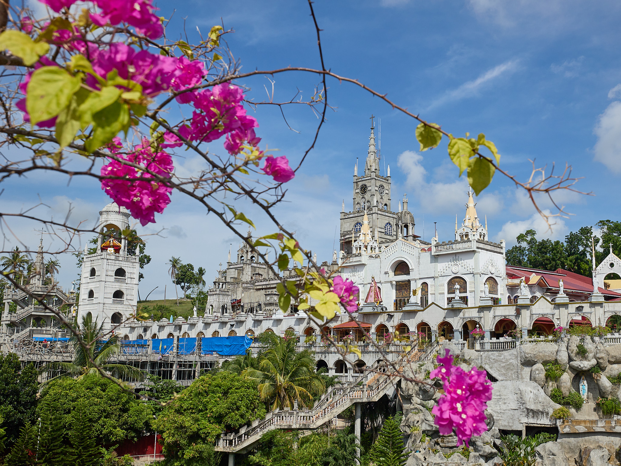 The white towers, spires and arcades of the Simala Shrine stacked up a hillside, seen through pink bougainvillea, with a clock tower at the centre, palm trees below and blue sheeting on scaffolding along one low wing