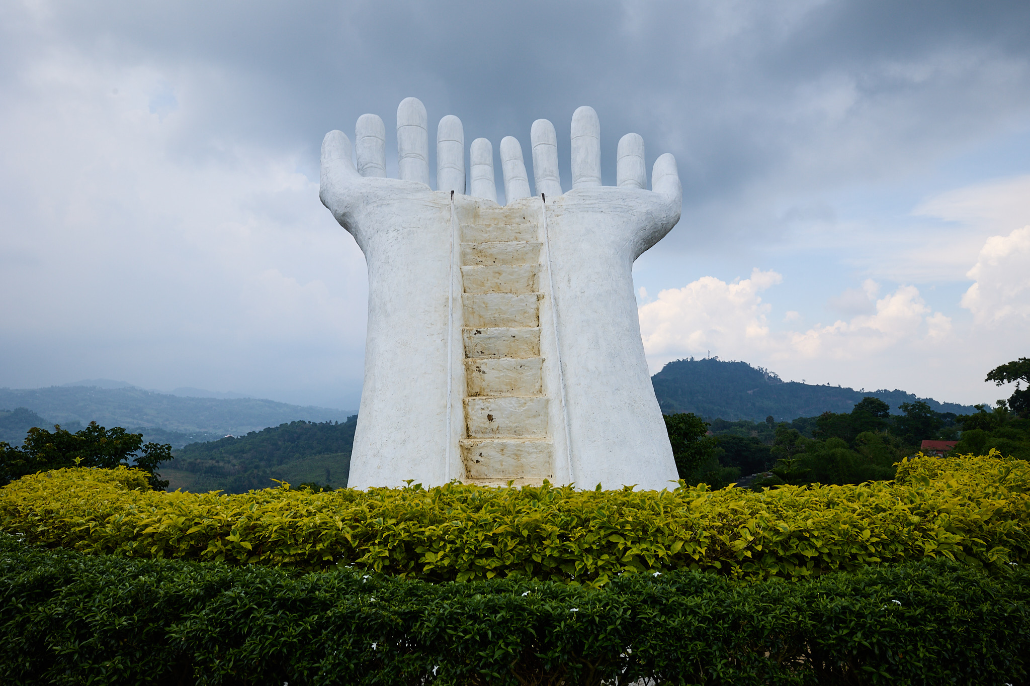 Two giant white hands rising side by side above a hedge, fingers curled upward, with a narrow staircase running up between them, under a cloudy sky with hills behind