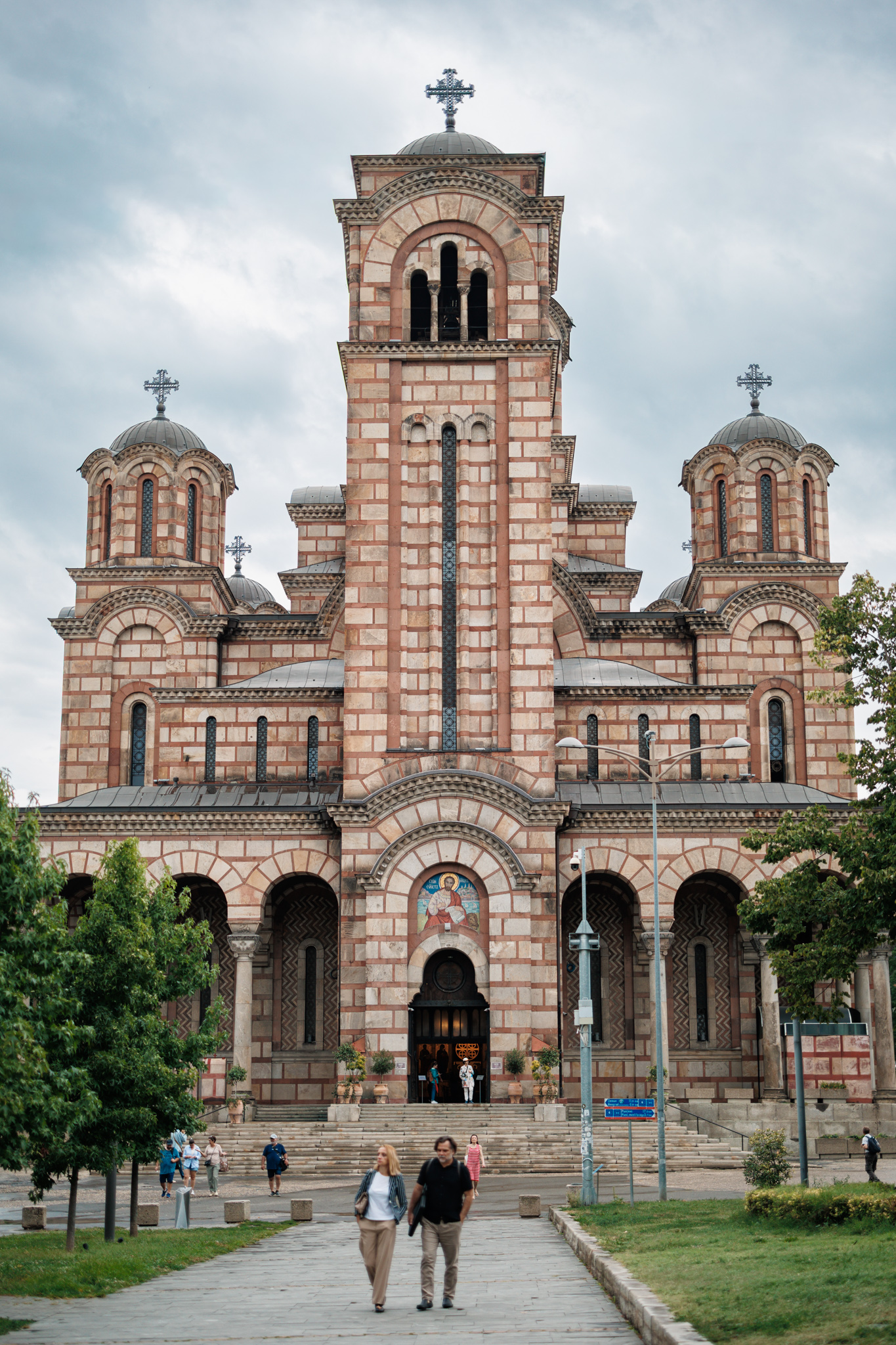 The front of St. Mark’s Church in Belgrade, with a tall bell tower between two domed towers, bands of red and pale stone, and two people walking on the path in front