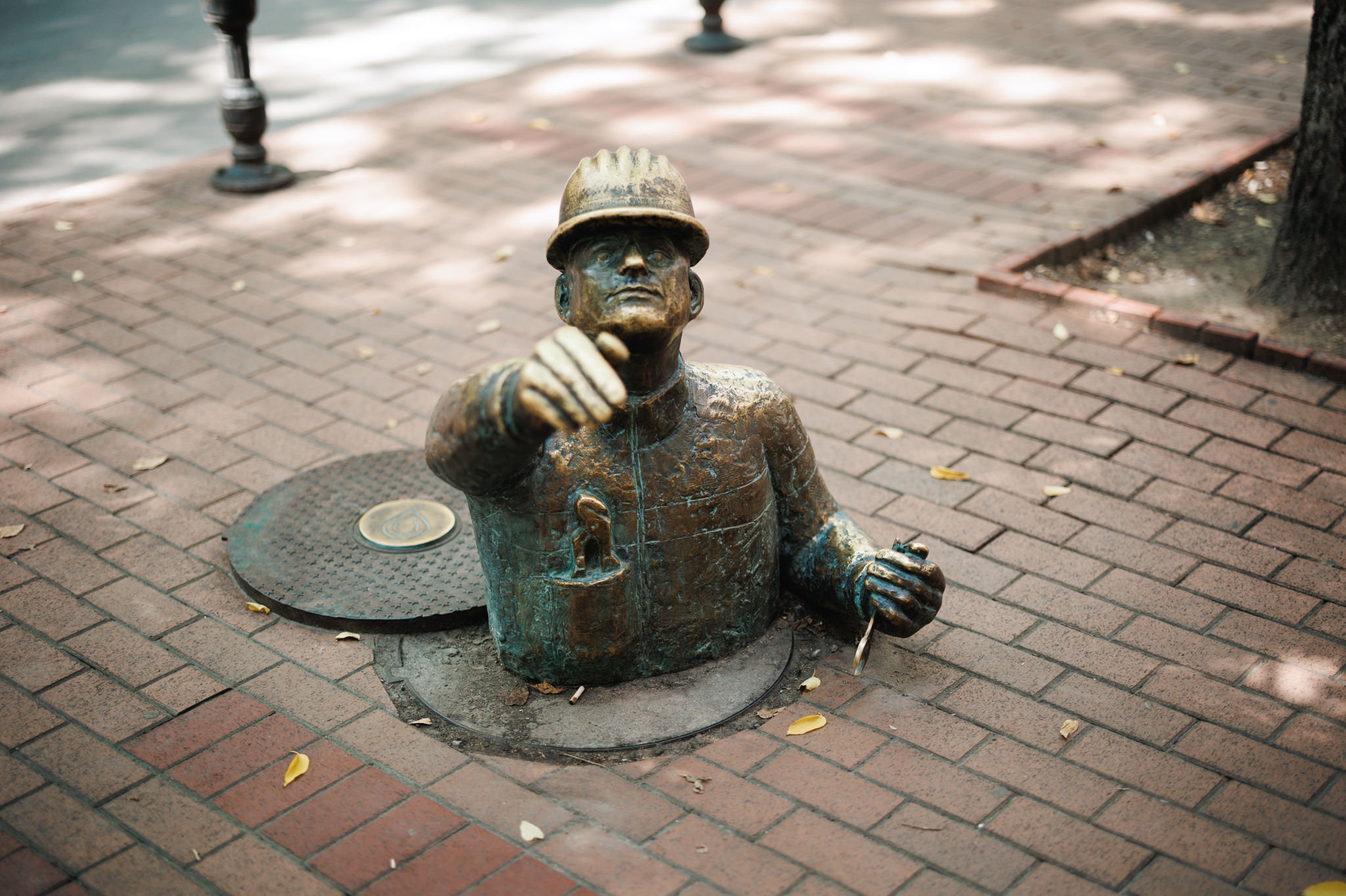 A bronze plumber in a hard hat emerging from a manhole in a brick pavement in Subotica, with one hand extended towards the viewer.
