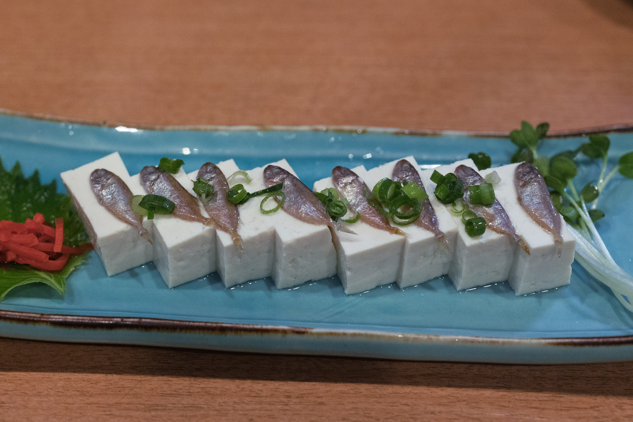 Sukugarasu tofu on a long light-blue plate: eight blocks of white tofu in a row, each topped with one small salted silver fish and sliced spring onion, with shiso, pickled red ginger and cress at the ends