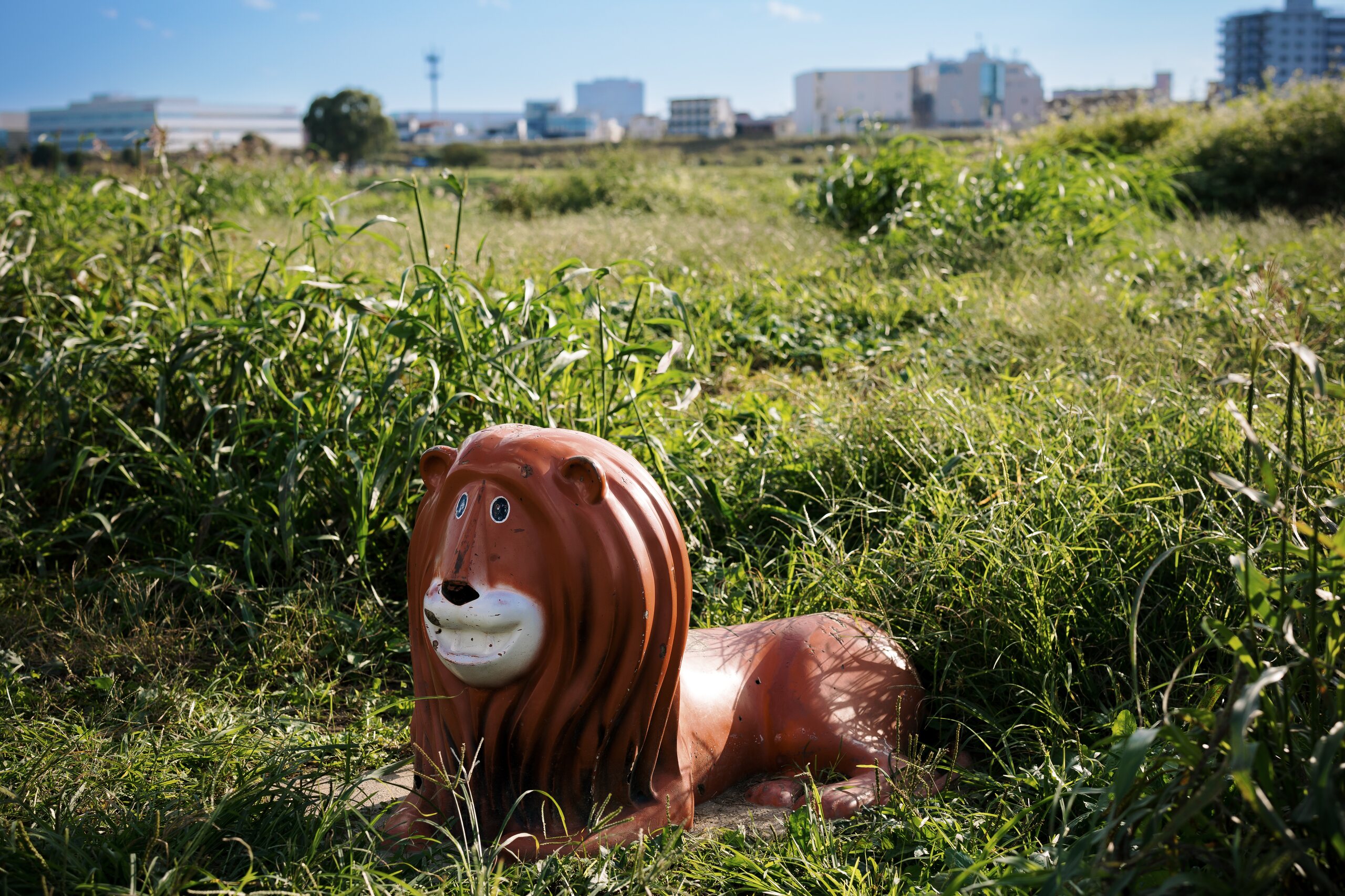 A weathered red playground lion lying in tall grass beside the Tama River in Tokyo.