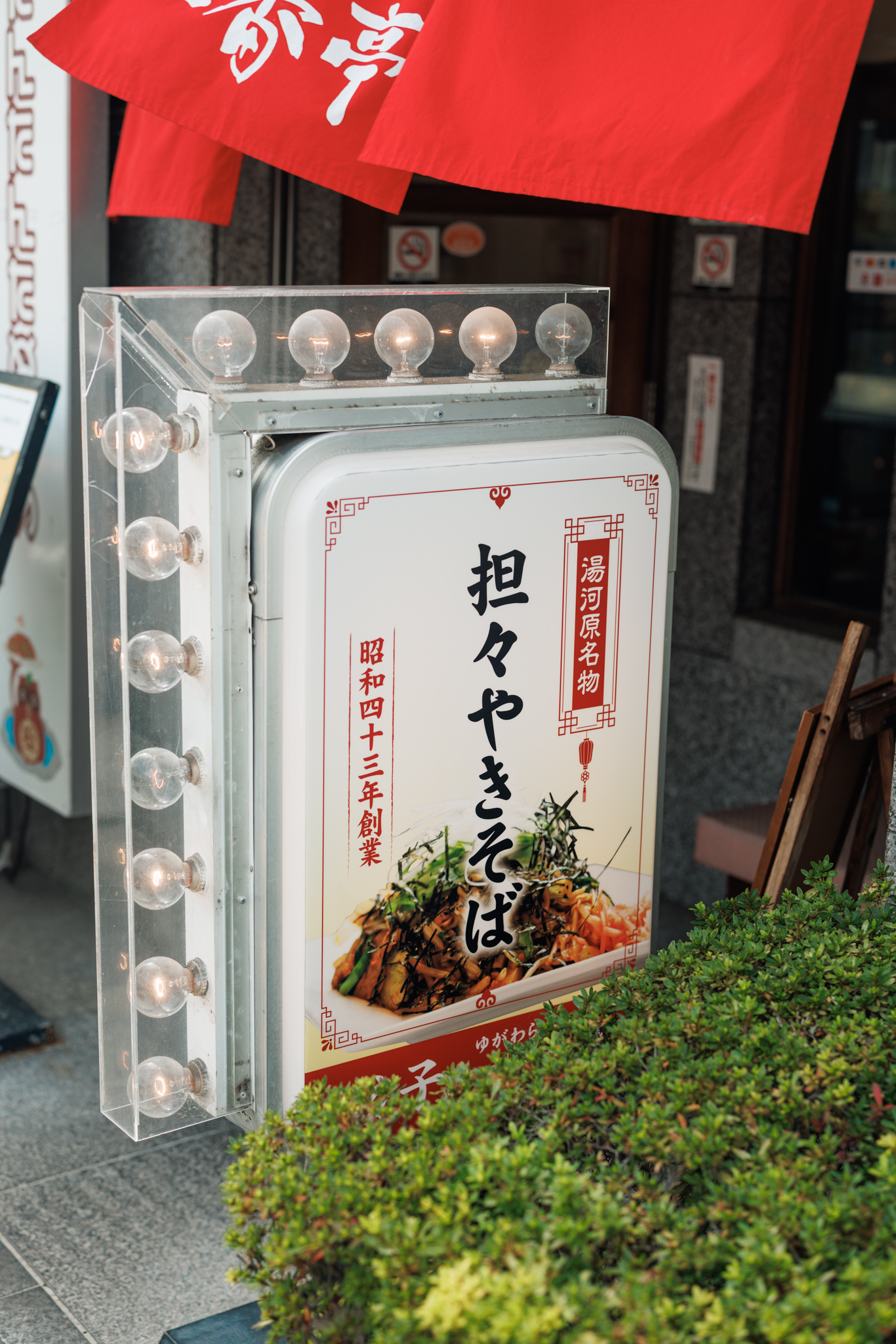 A free-standing sign framed with clear light bulbs, advertising tantan yakisoba as a Yugawara speciality with a photo of the dish and the founding year Shōwa 43, beside a shrub and under a red noren curtain