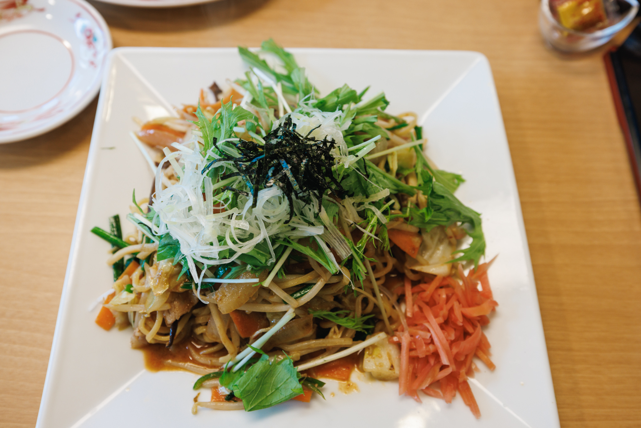 The same plate of tantan yakisoba seen from above: a mound of mizuna and shredded leek with nori in the centre, red pickled ginger at the side and sauce at the edges