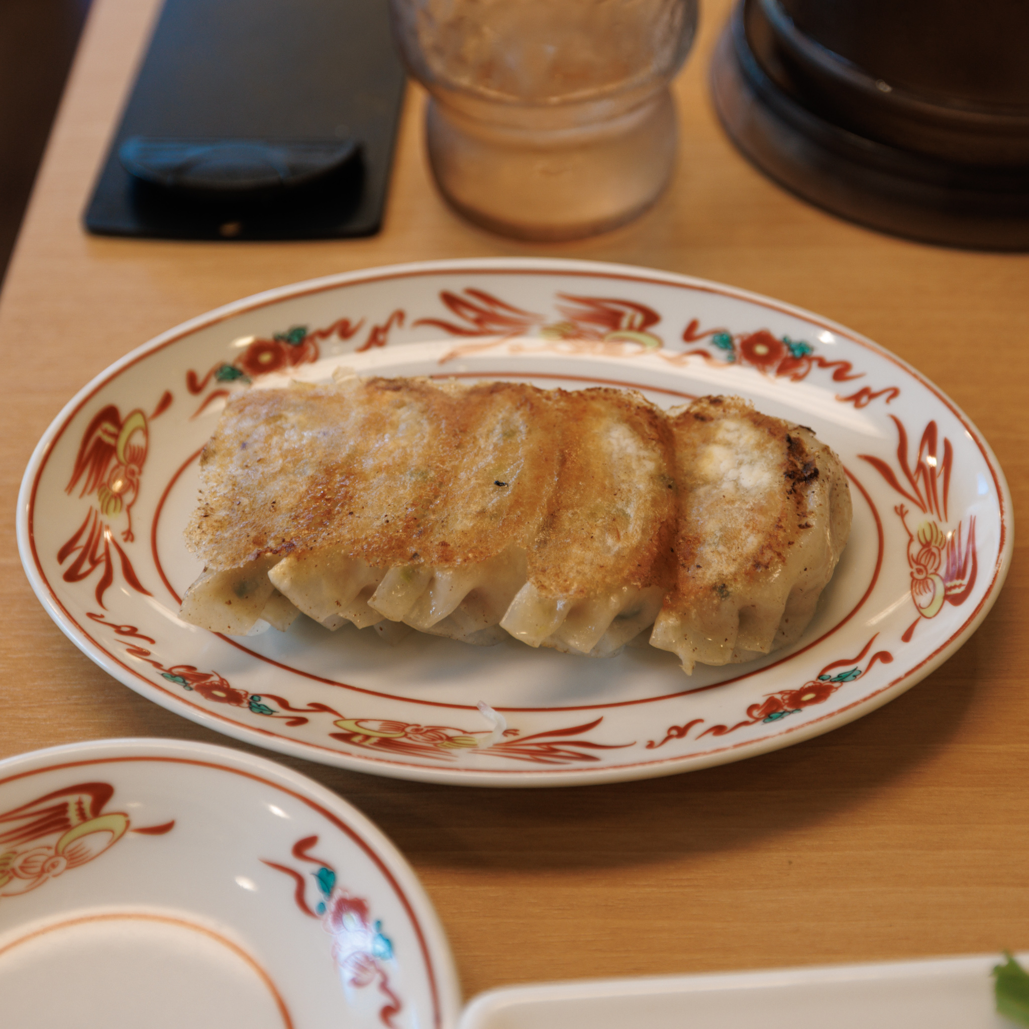 Five pan-fried gyoza joined by a browned crust, on an oval plate with a red phoenix-and-flower border