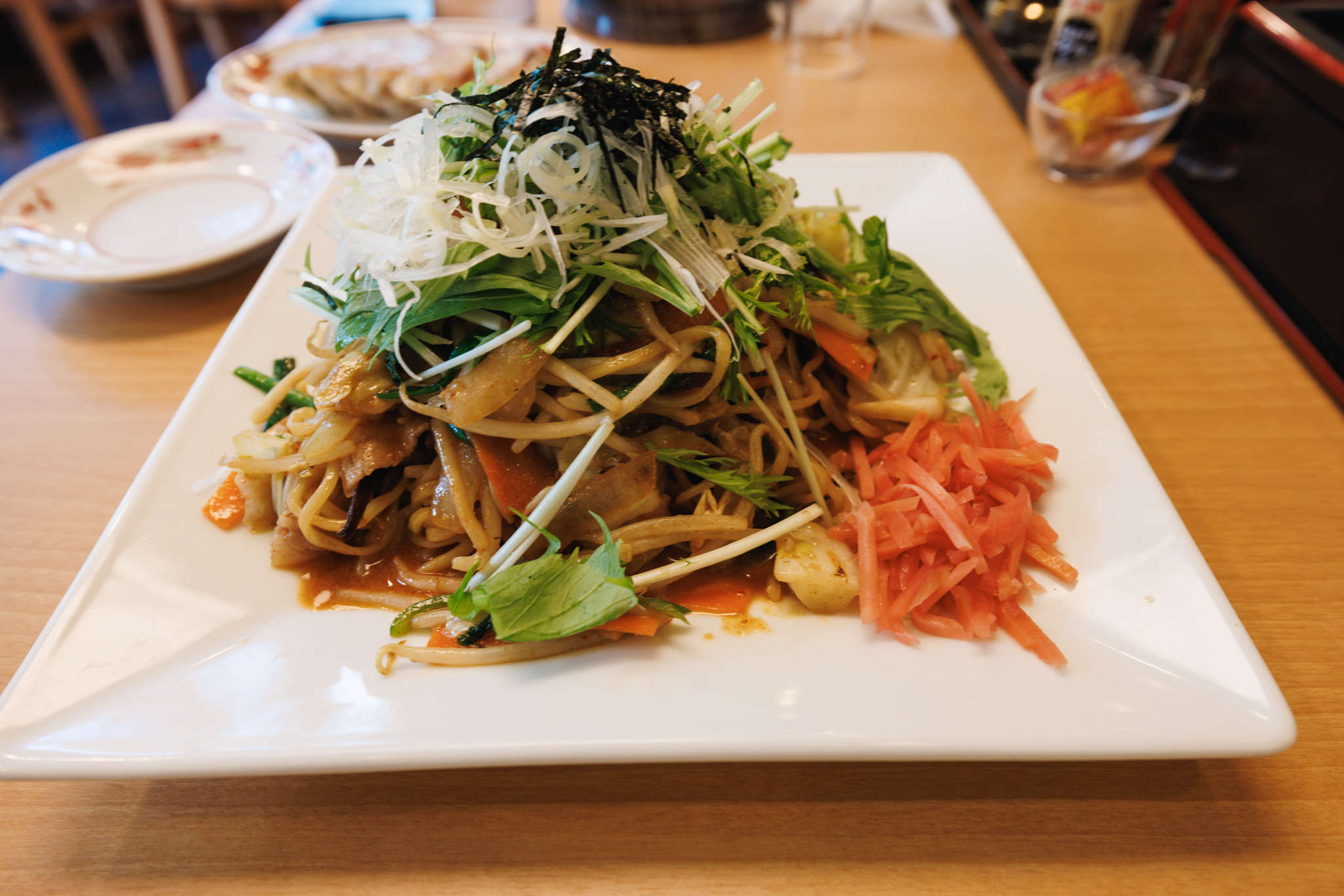 A hill of tantan yakisoba on a square white plate, topped with mizuna, finely shredded white leek and shredded nori, with a heap of red pickled ginger at one side and reddish-brown sauce on the plate; a plate of gyoza behind