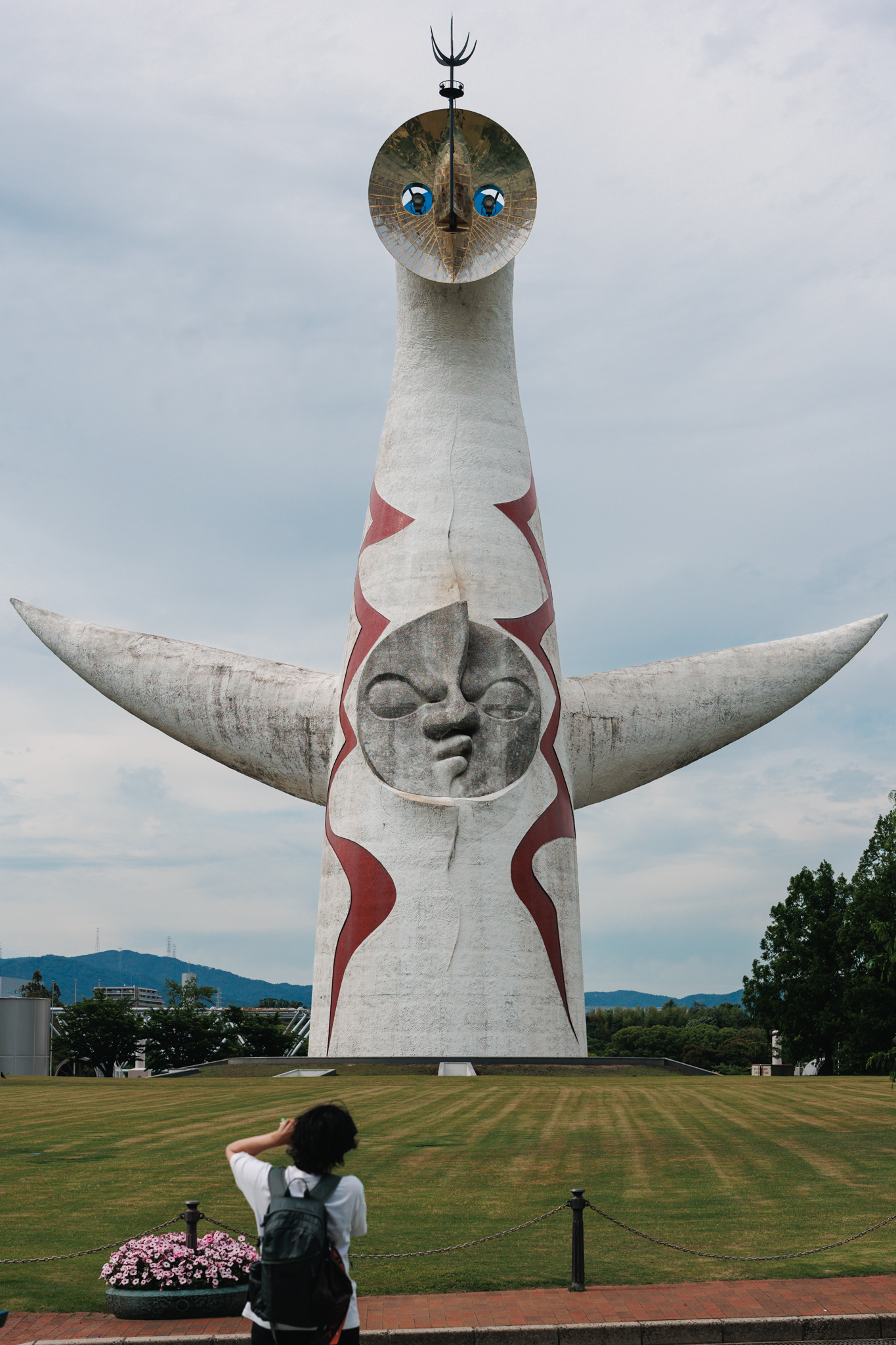 The Tower of the Sun seen from the front across a lawn, with its golden face on top, a grey sun face on its body and outstretched arms, and a visitor photographing it in the foreground