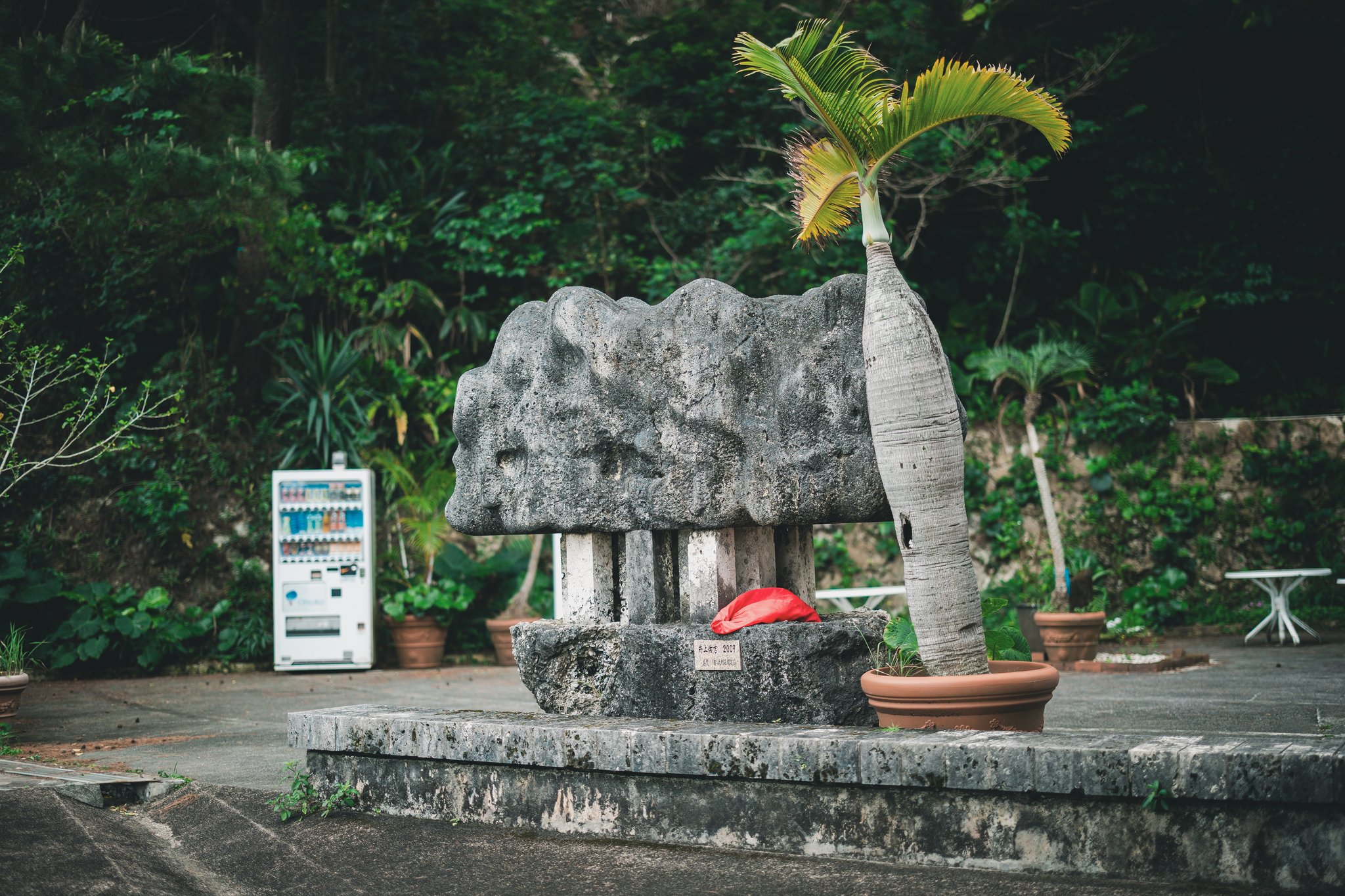 A massive grey stone sculpture resting on stone posts, with a red cloth on its base, a potted palm beside it and a drinks vending machine behind