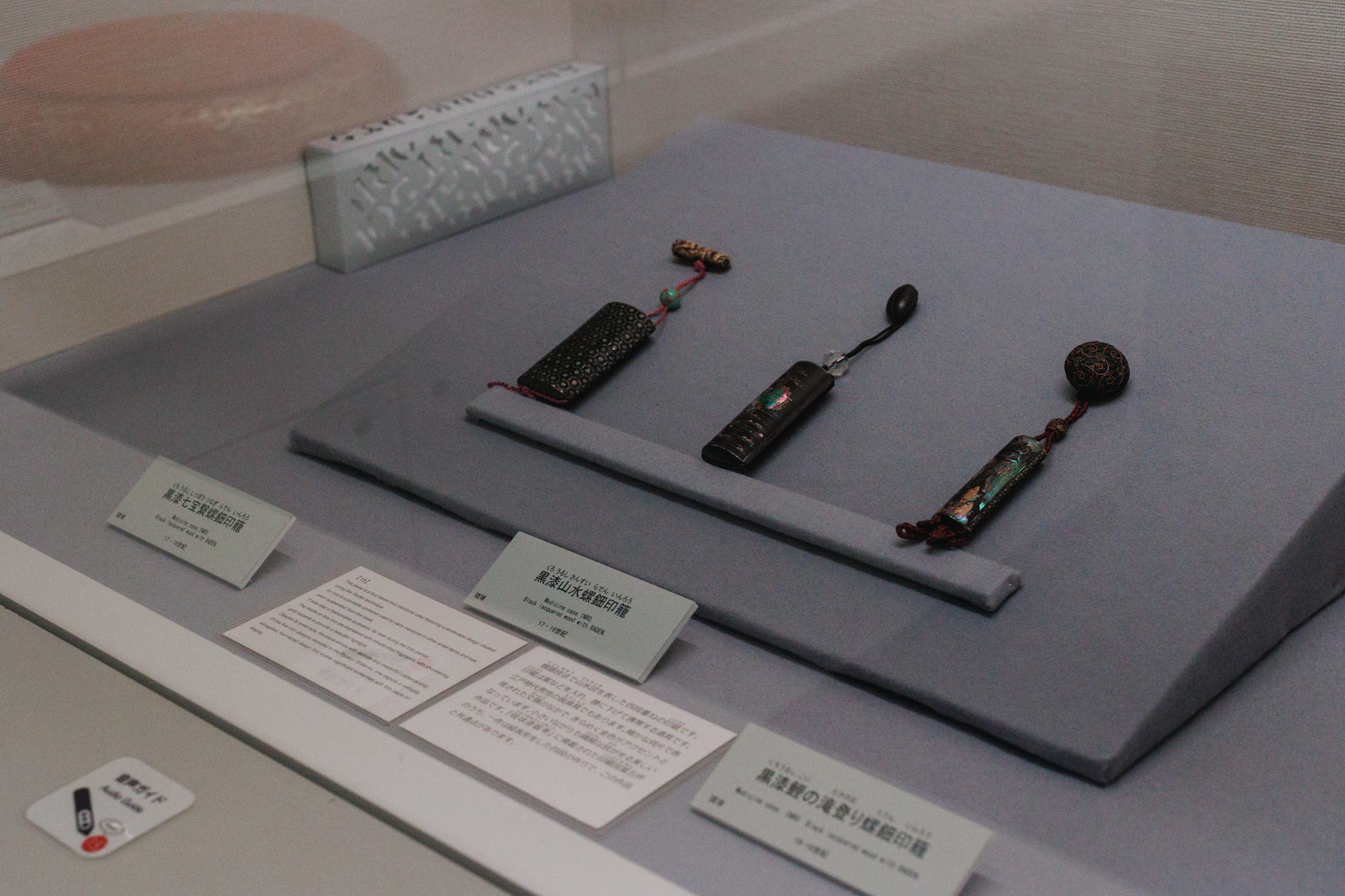 Three black lacquer inro inlaid with mother-of-pearl, with cords and beads, displayed on a grey stand with Japanese and English labels