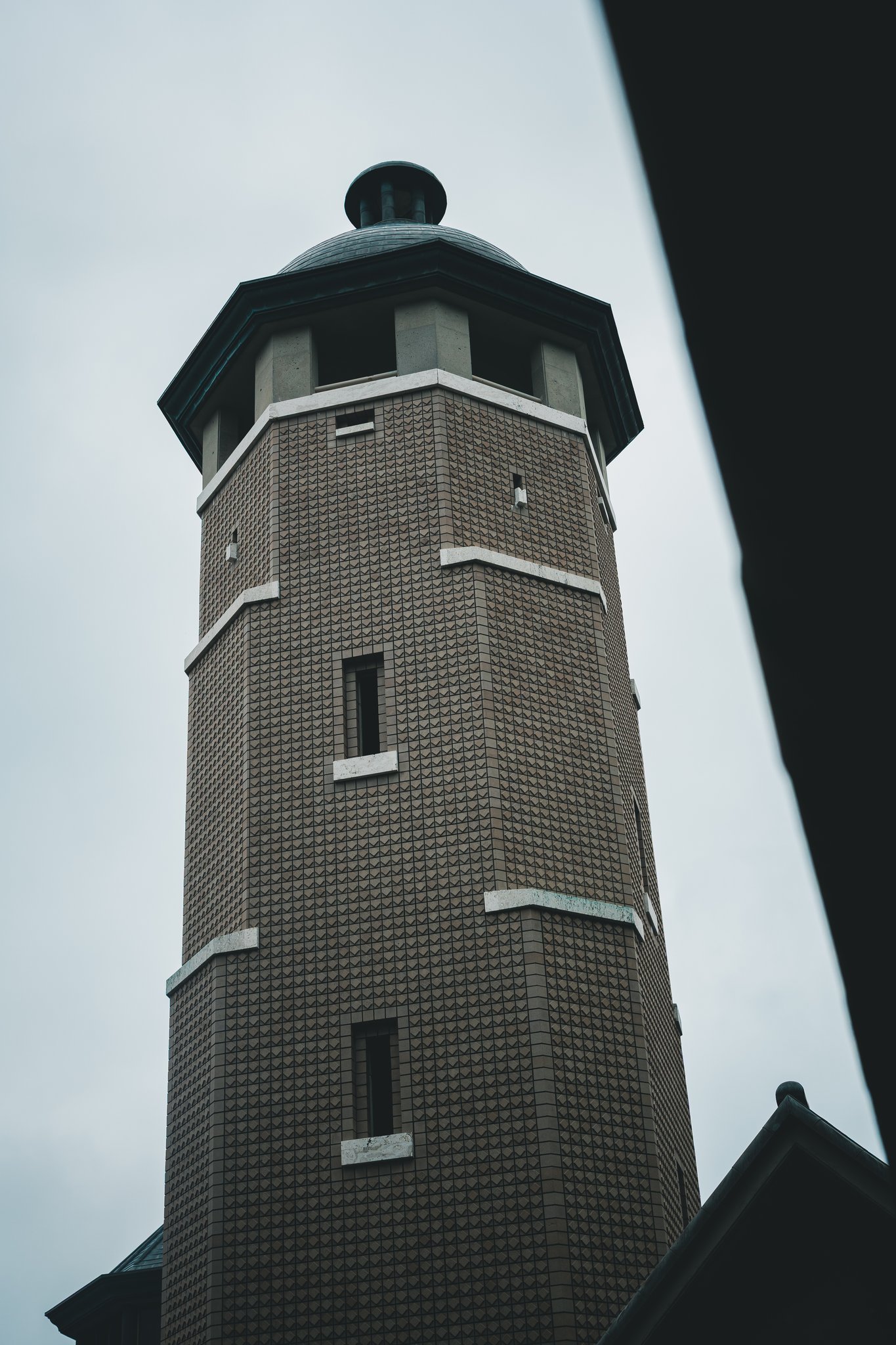 An octagonal tower with textured brown tiles, white bands, narrow windows, an open lookout and a dark dome against a grey sky