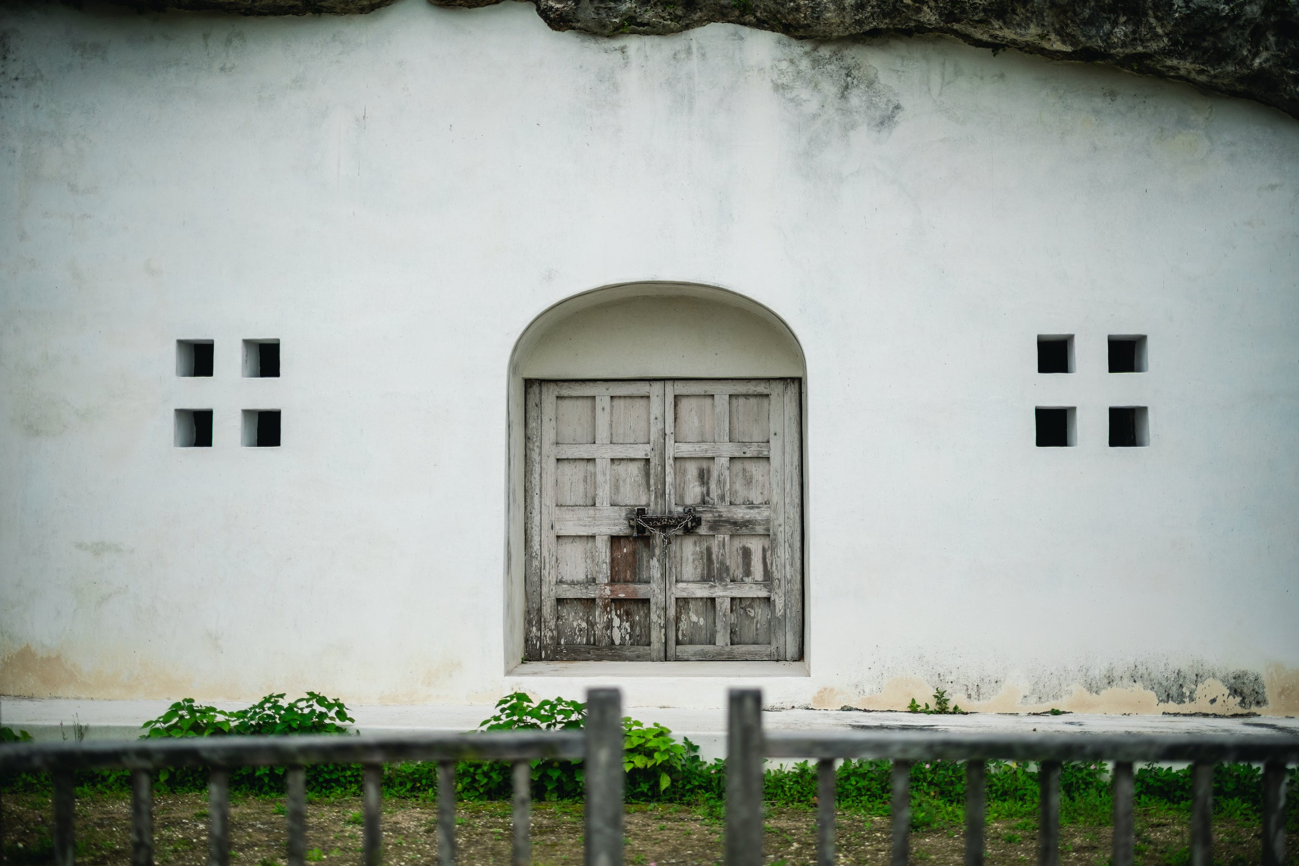 The chained and padlocked wooden double door of the west chamber of Urasoe Yōdore, set in a white wall under overhanging rock, with small square windows on each side and a fence in the foreground