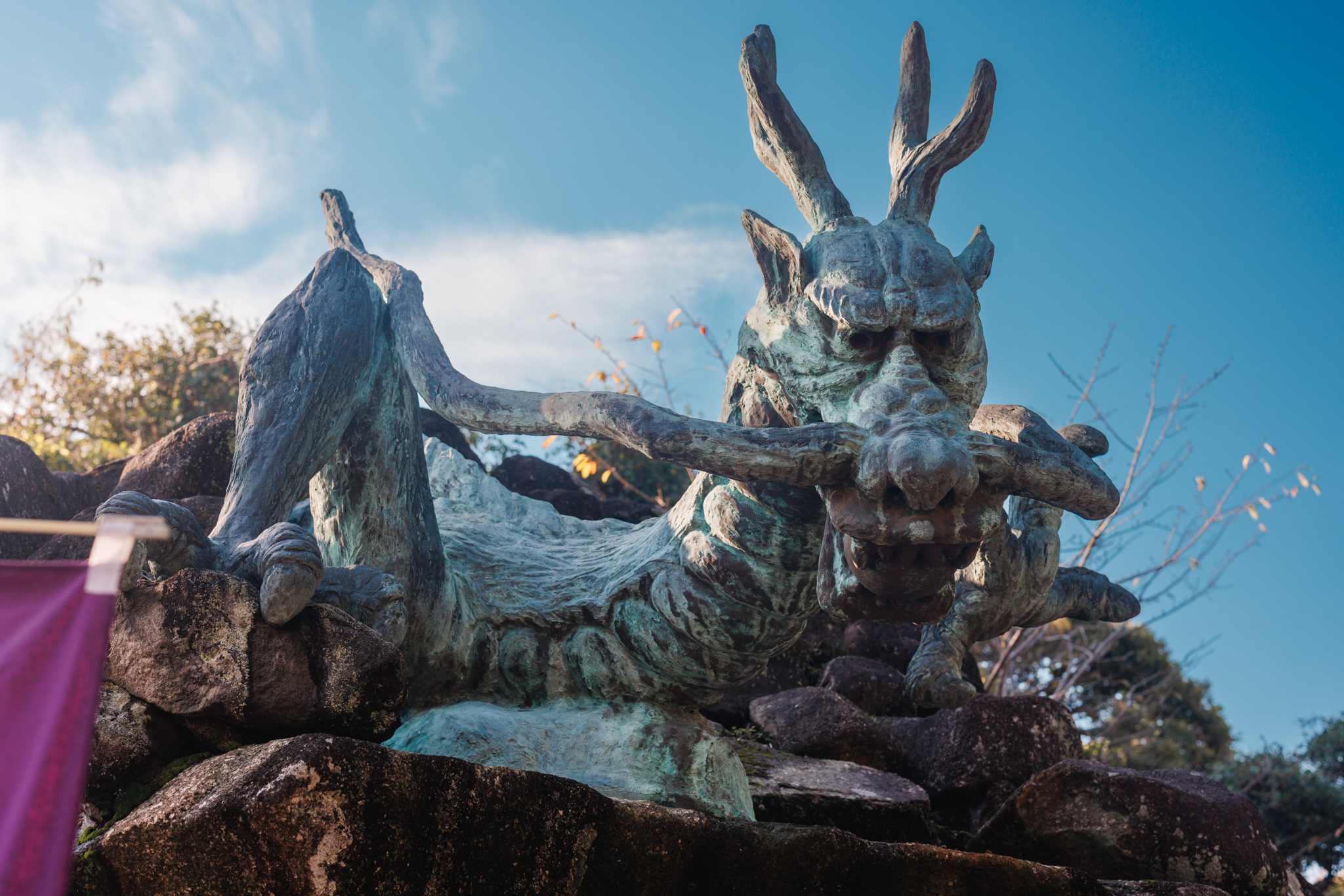 A weathered bronze dragon with antler-like horns and long whiskers, seen from below against a blue sky, its claws resting on rocks