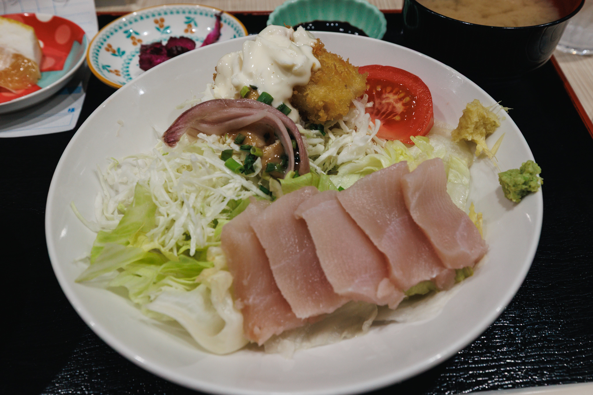 A white plate of shark sashimi: five thick, pale pink slices laid over lettuce and shredded cabbage, with grated ginger and wasabi at the side, a fried piece under tartar sauce, a slice of tomato and a small vinegared dish topped with spring onion behind