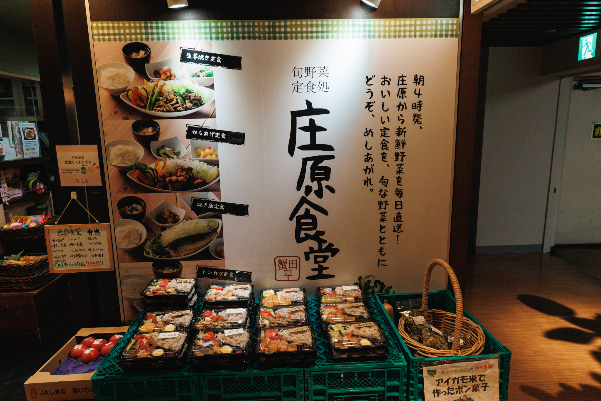 The entrance of Shobara Shokudo: a large sign with the restaurant’s name in brush lettering, a message about vegetables leaving Shobara at 4 a.m., photos of set meals, and crates of packed lunch boxes, tomatoes and bags of puffed rice in front