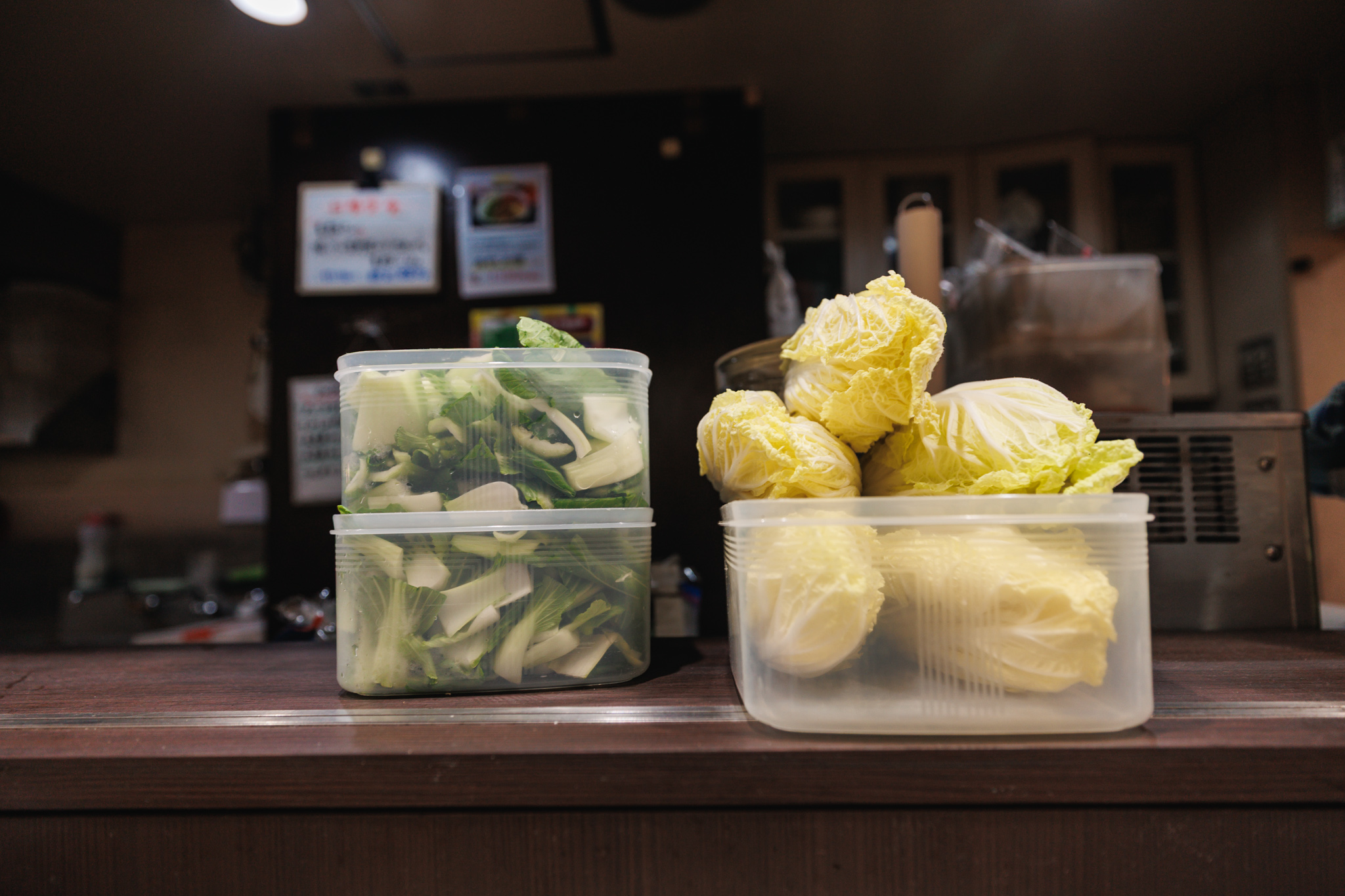 Two clear plastic containers on a wooden counter, one stacked with cut green leafy vegetables and the other with heads of Chinese cabbage, in front of a dimly lit kitchen
